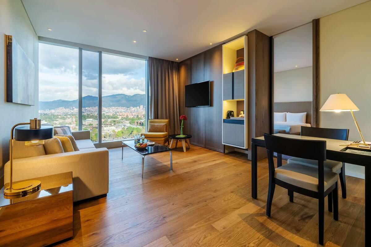 Ein modernes Hotelzimmer mit großem Fenster und Blick auf die Stadt, einer Couch, einem Esstisch und einem an der Wand montierten Fernseher.