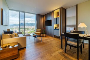 Una habitación de hotel moderna con un amplio ventanal que ofrece vistas a la ciudad, un sofá, una mesa de comedor y un televisor en la pared.