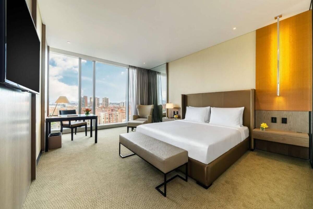 Une chambre d’hôtel avec un grand lit, un bureau, une chaise, un banc et une grande fenêtre donnant sur la ville.