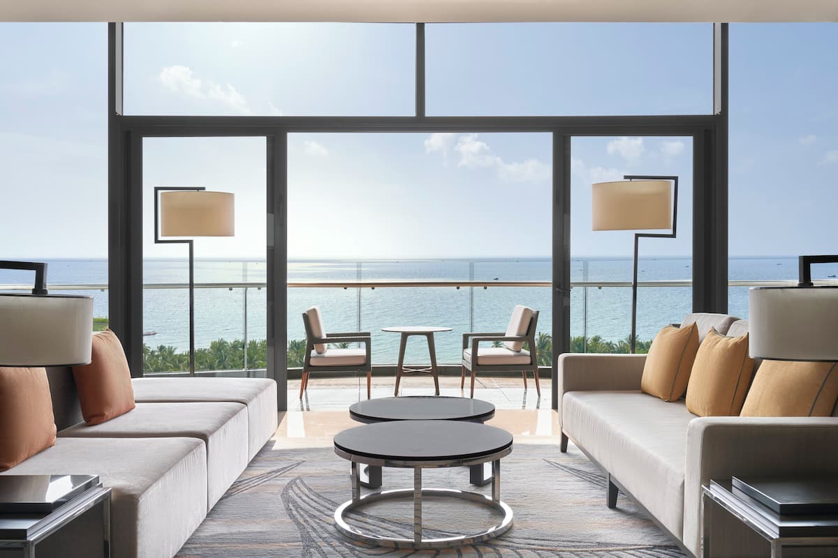 Una habitación de hotel moderna con un balcón grande, dos sofás, una mesa redonda y dos lámparas.