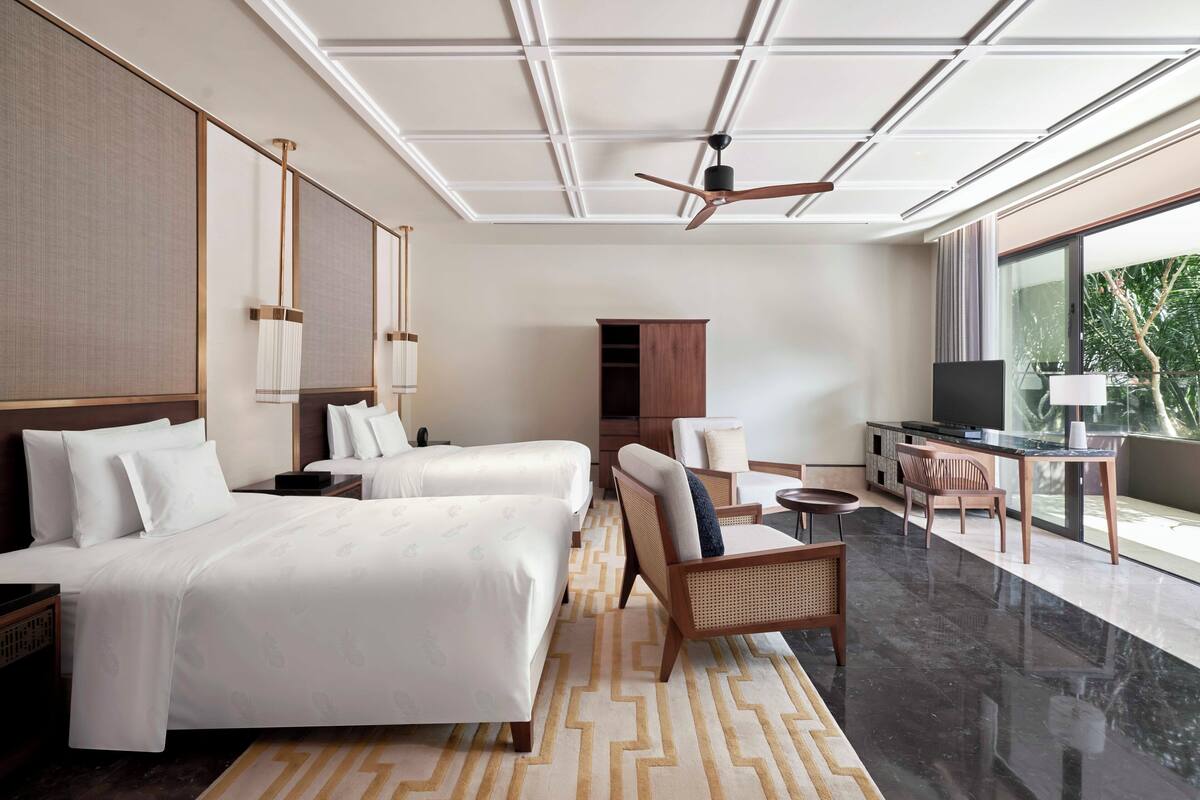 A modern hotel room with two beds, a seating area, a television, and a large window offering a view of greenery.