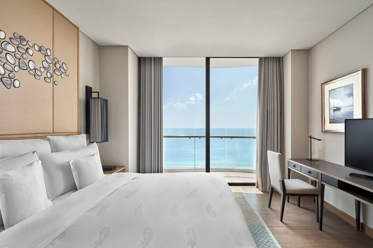 A modern hotel room with a large bed, a desk, and a view of the ocean through a large window.