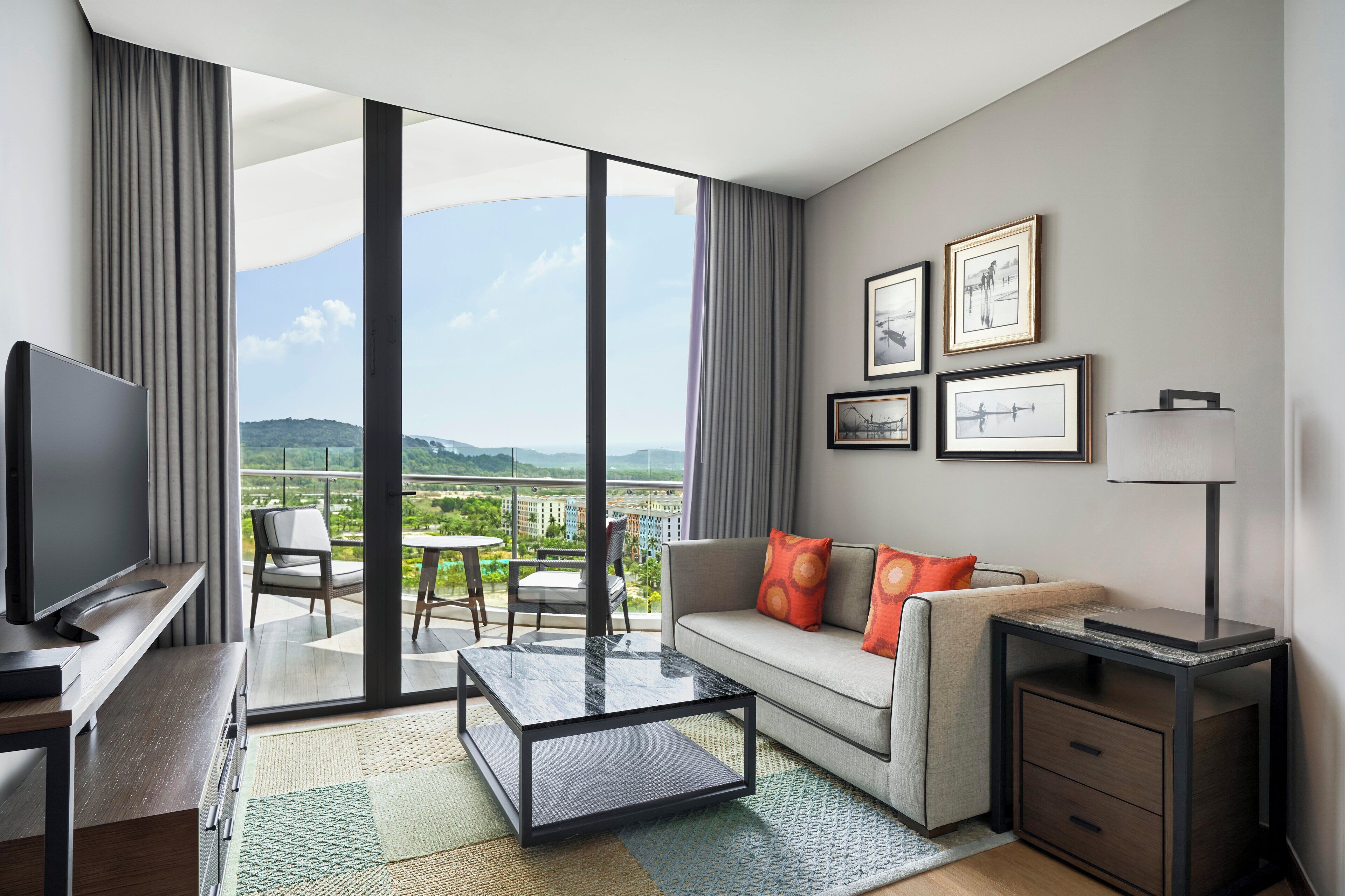 Une chambre d’hôtel moderne dotée d’un grand balcon, d’un canapé avec des coussins orange, d’un téléviseur à écran plat, d’un bureau avec une lampe et de tableaux encadrés au mur.