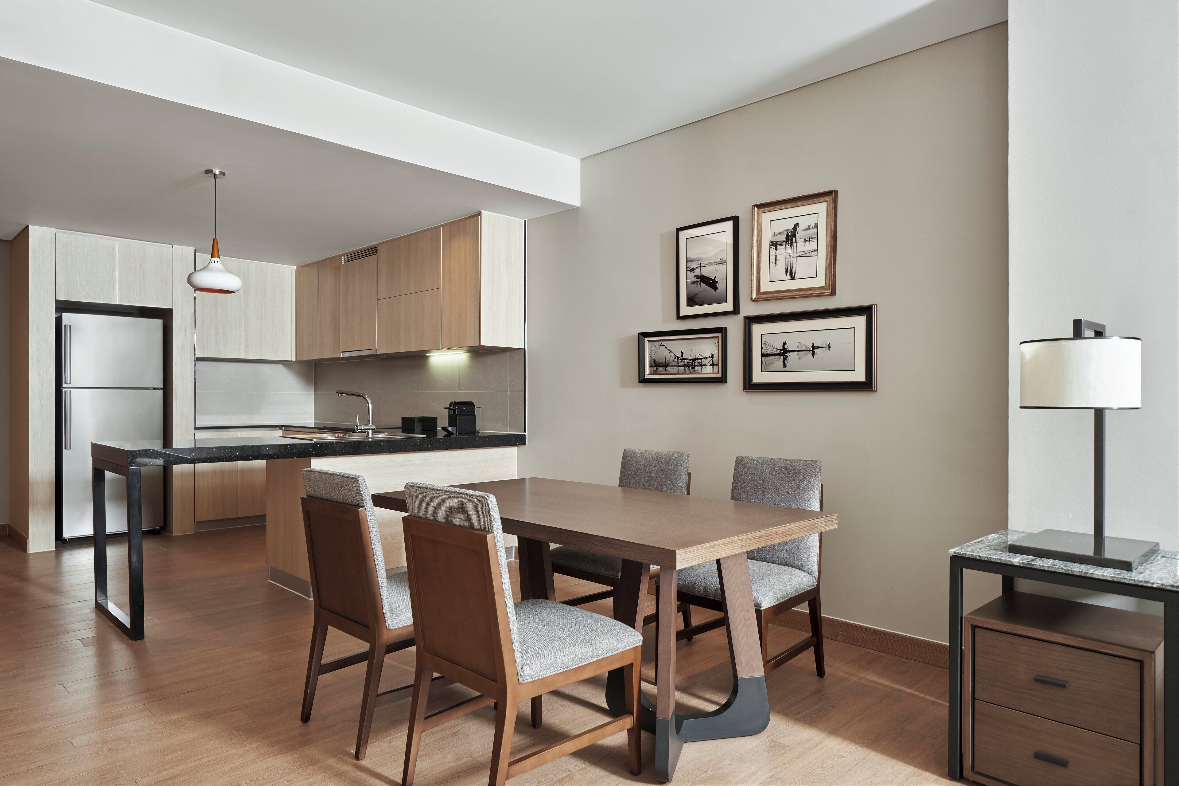 Cozinha moderna com armários de madeira, uma ilha integrada e um espaço de refeições com mesa e cadeiras. Há quadros nas paredes e um abajur sobre uma mesinha.