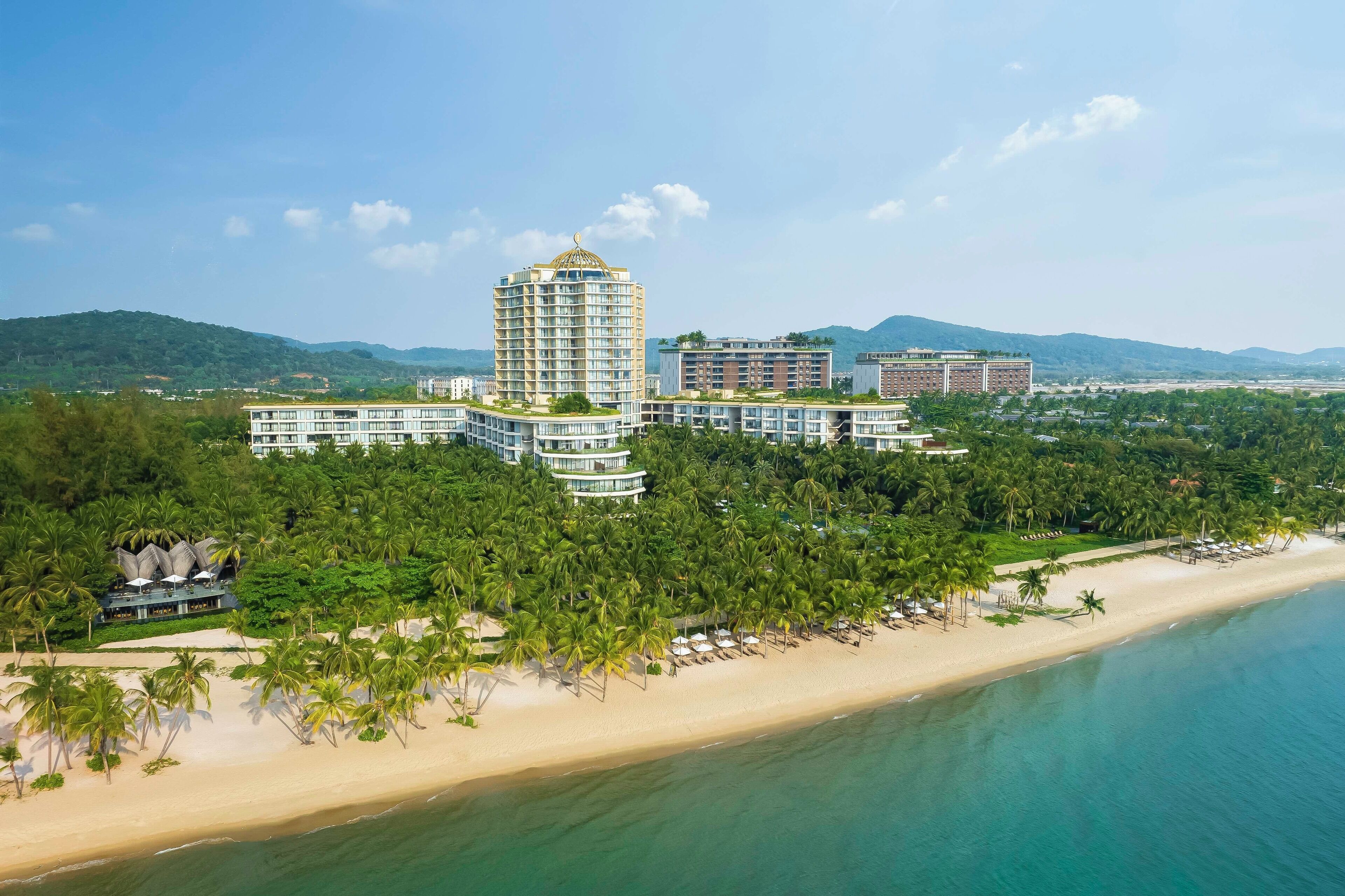 A coastal hotel with a central tower, multiple buildings, and a beach with palm trees.