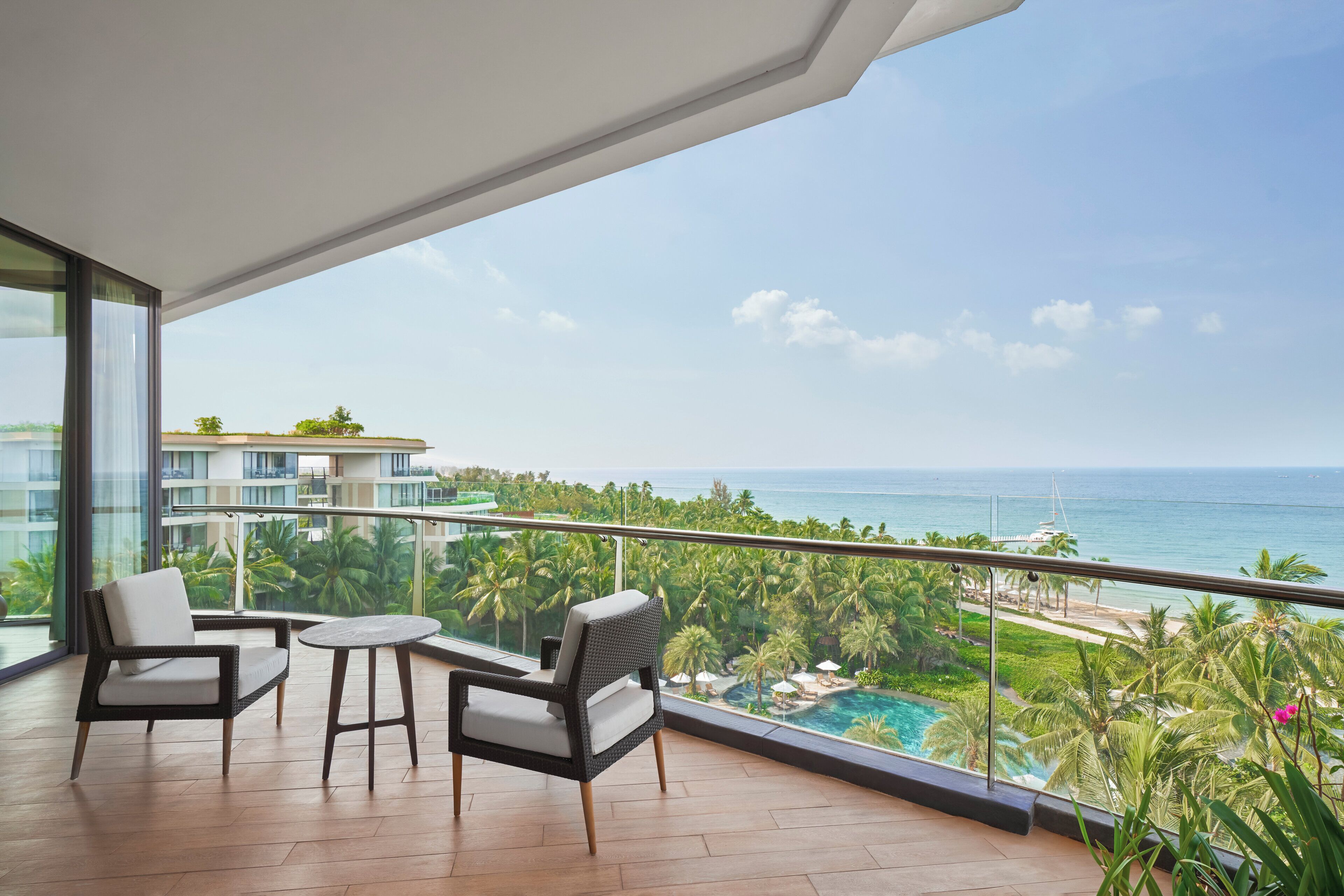 Un balcon avec des fauteuils en osier et une petite table, offrant une vue sur une piscine et l’océan.