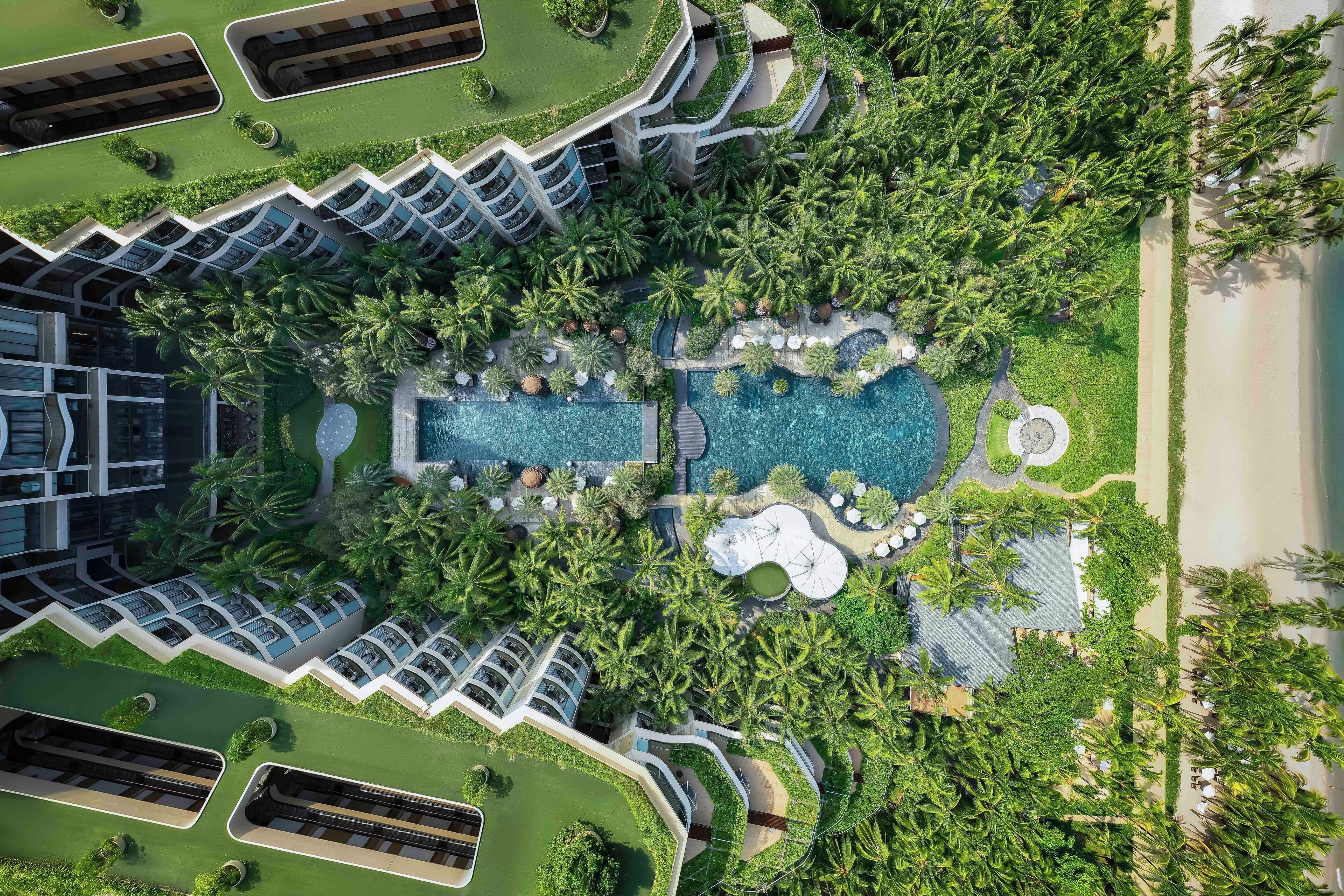An aerial view of a resort with multiple swimming pools, lush greenery, and a beach area.