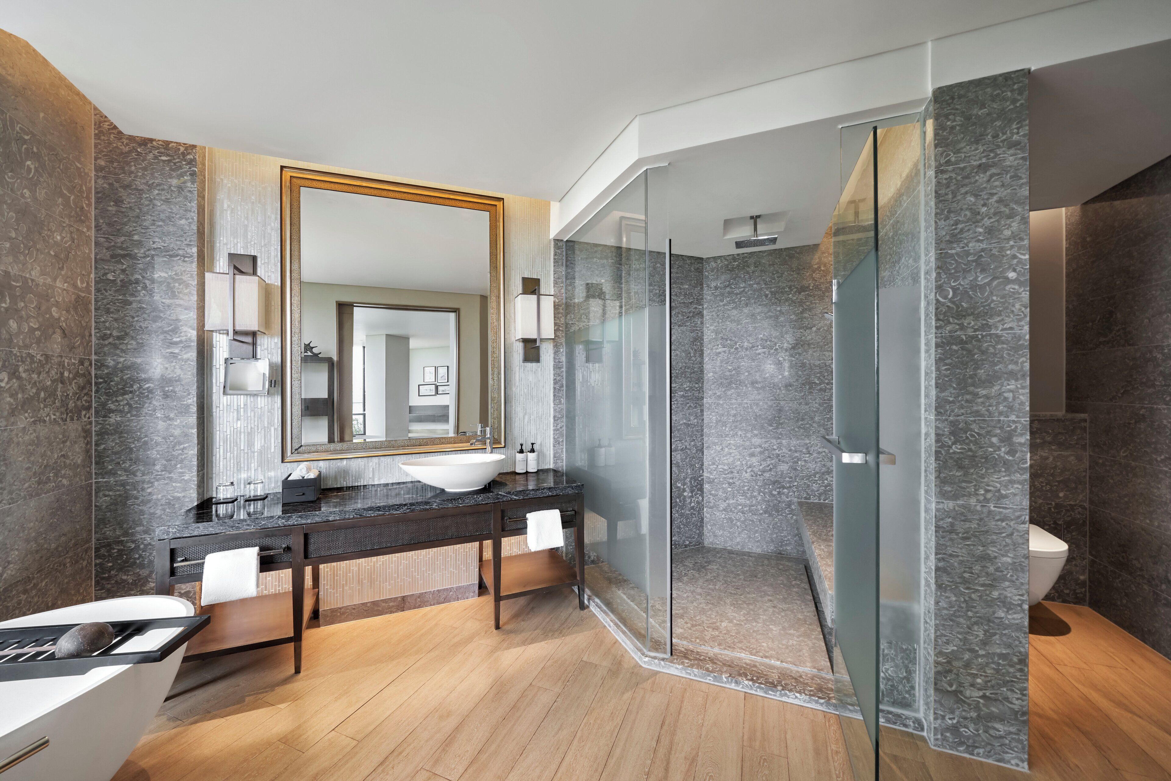 A modern bathroom with a double vanity, a large mirror, a glass-enclosed shower, and marble walls.
