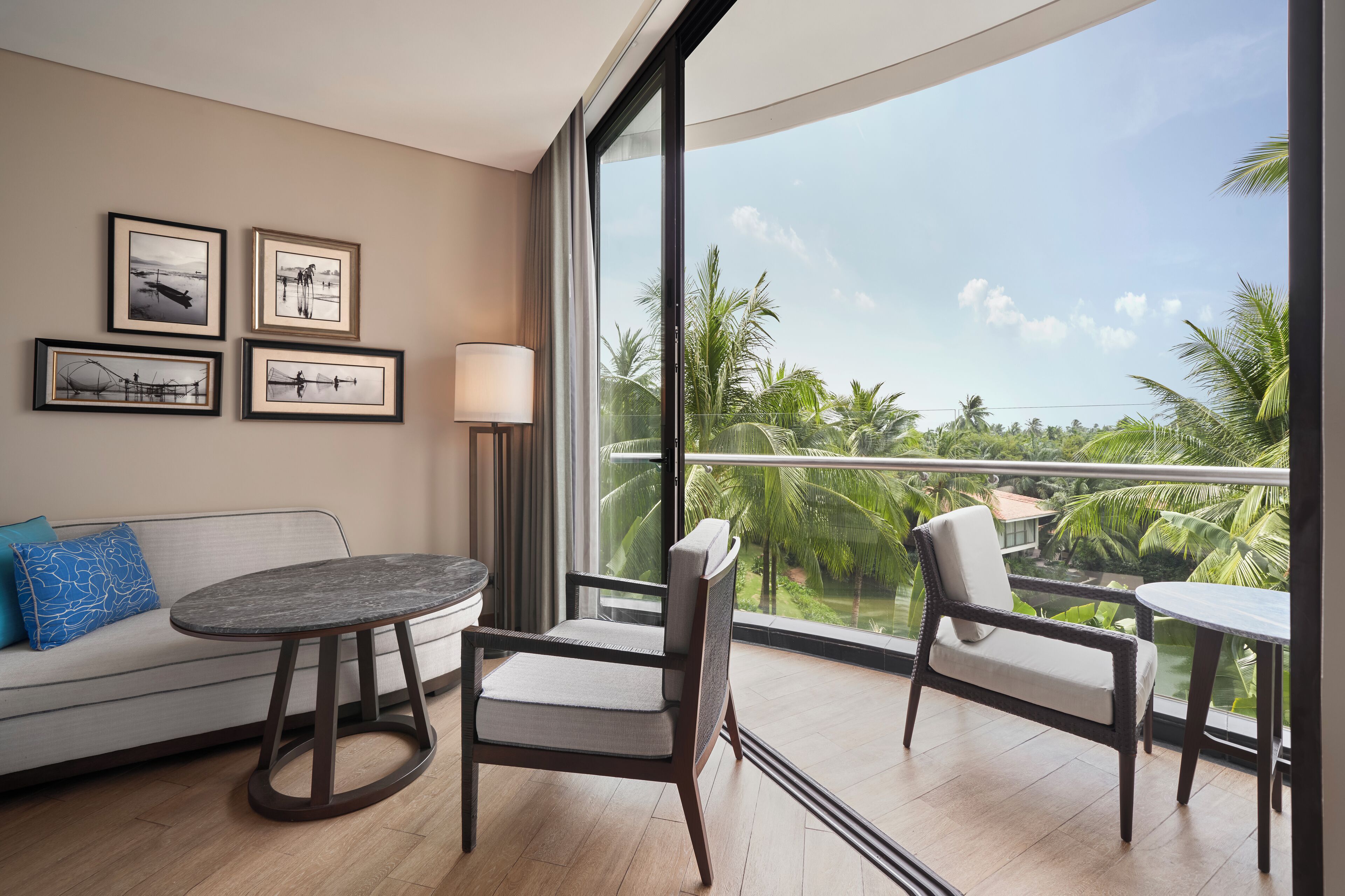 A modern hotel room with a balcony, a sofa with cushions, a round table, and framed artwork on the wall.