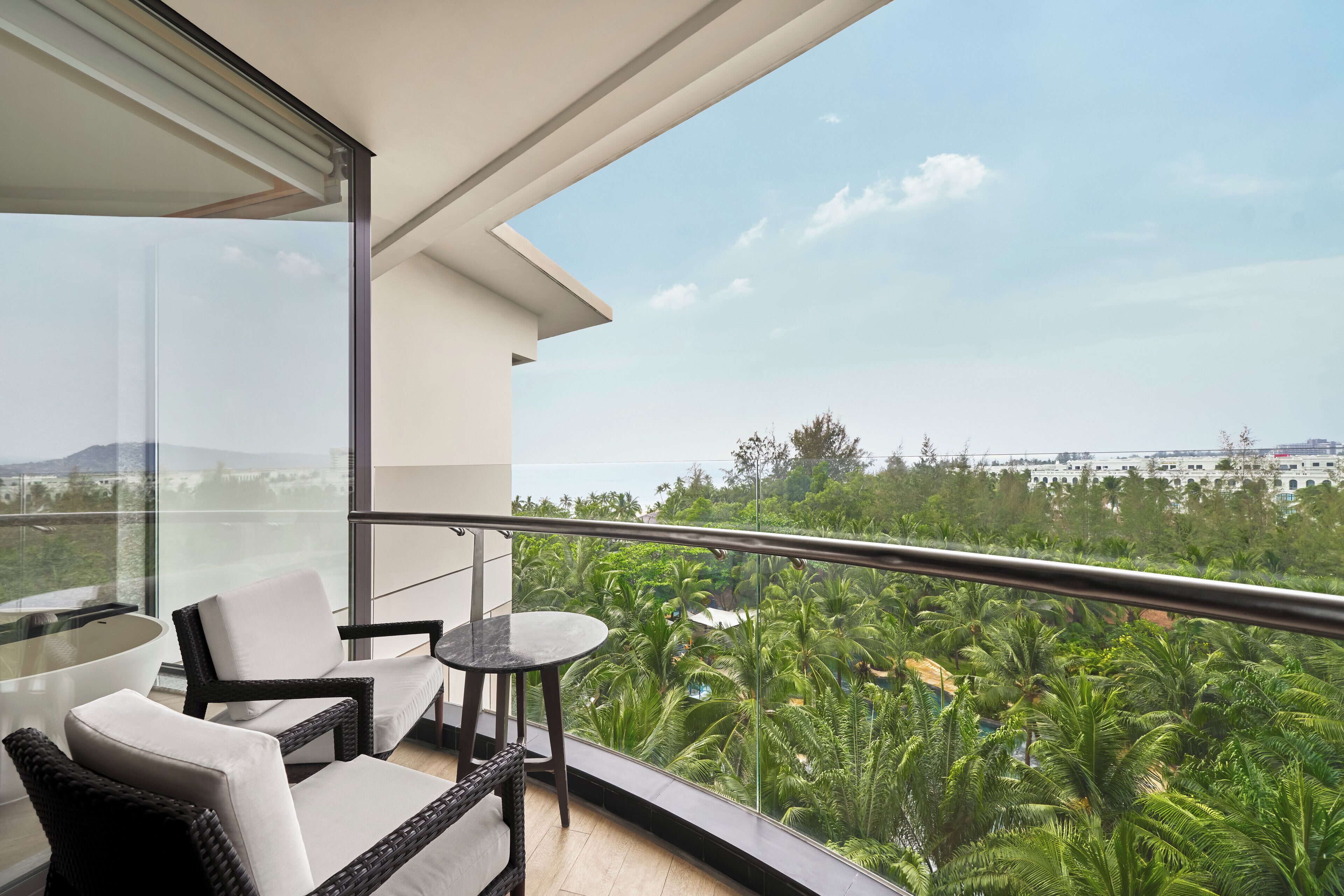 A balcony with wicker furniture, a glass table, and a view of lush greenery and distant buildings.
