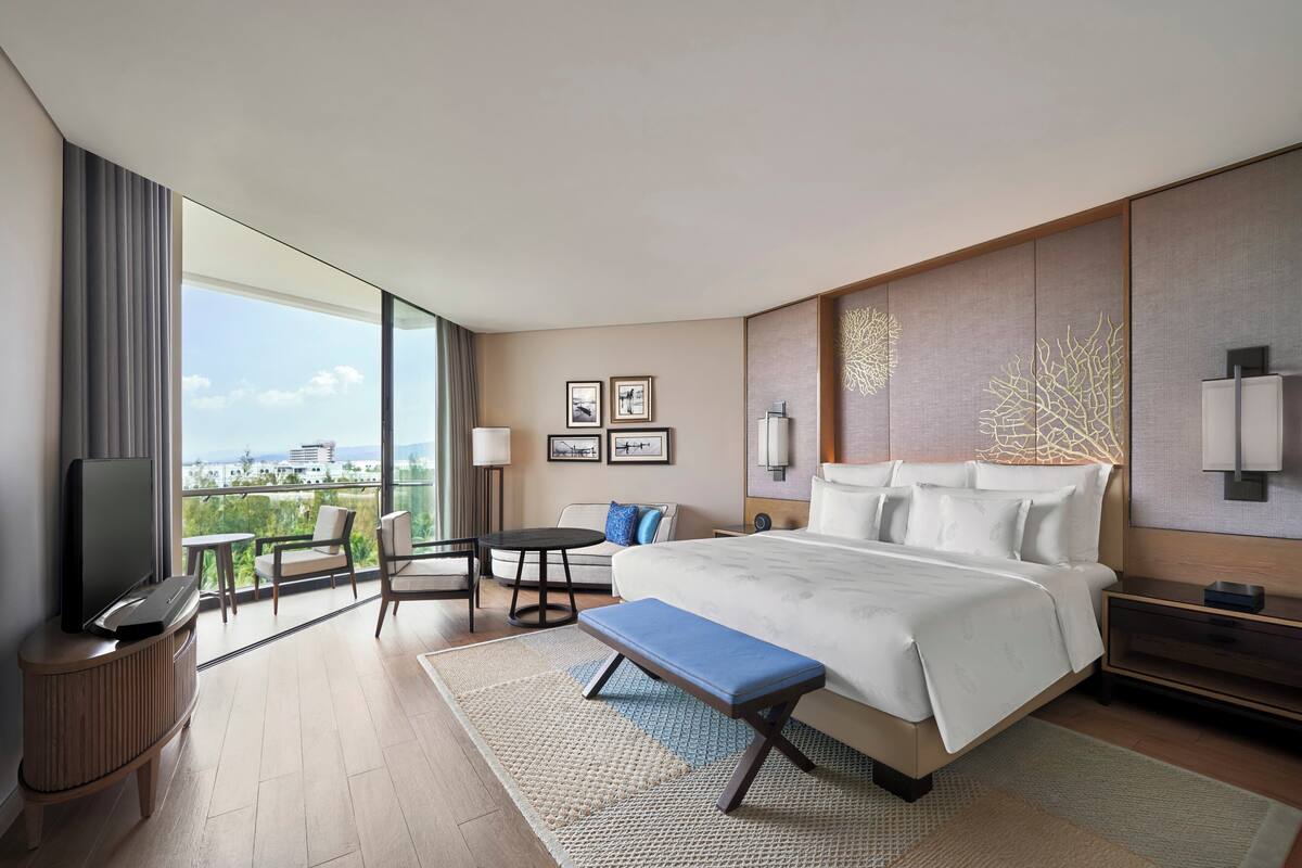 A modern hotel room with a large bed, a TV, a balcony with a view, and a seating area with a table and chairs.