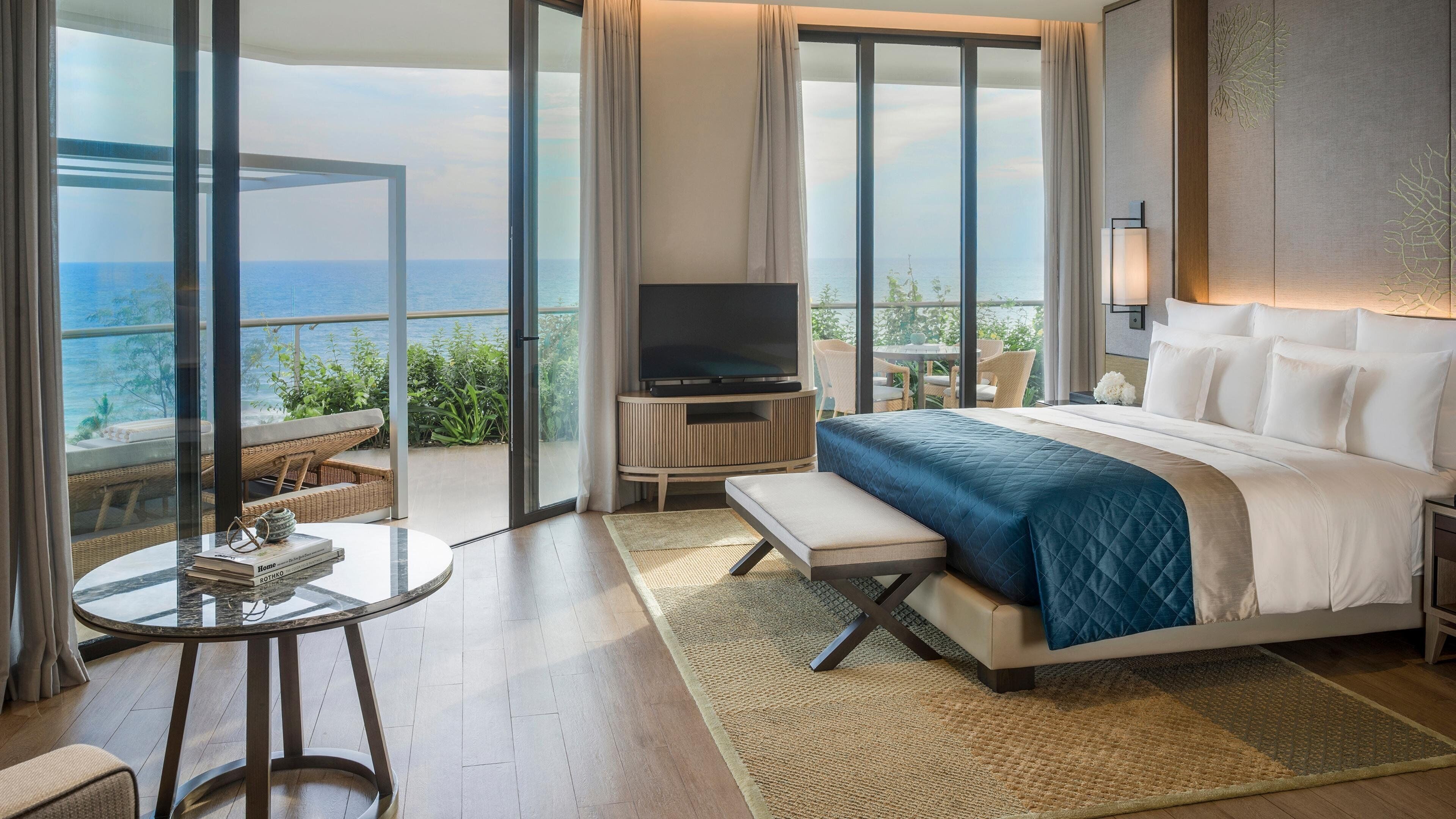 A hotel room with a large bed, a TV, a balcony with a view of the sea, and a dining area with a table and chairs.