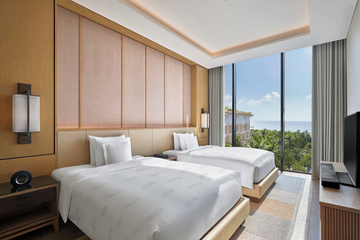 Habitación de hotel con dos camas, un televisor y un ventanal con vista al océano.
