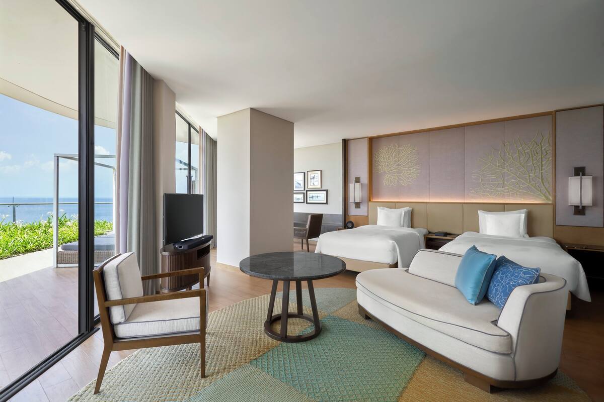 A modern hotel room with a large bed, a sofa, a TV, and a balcony with a view of the ocean.