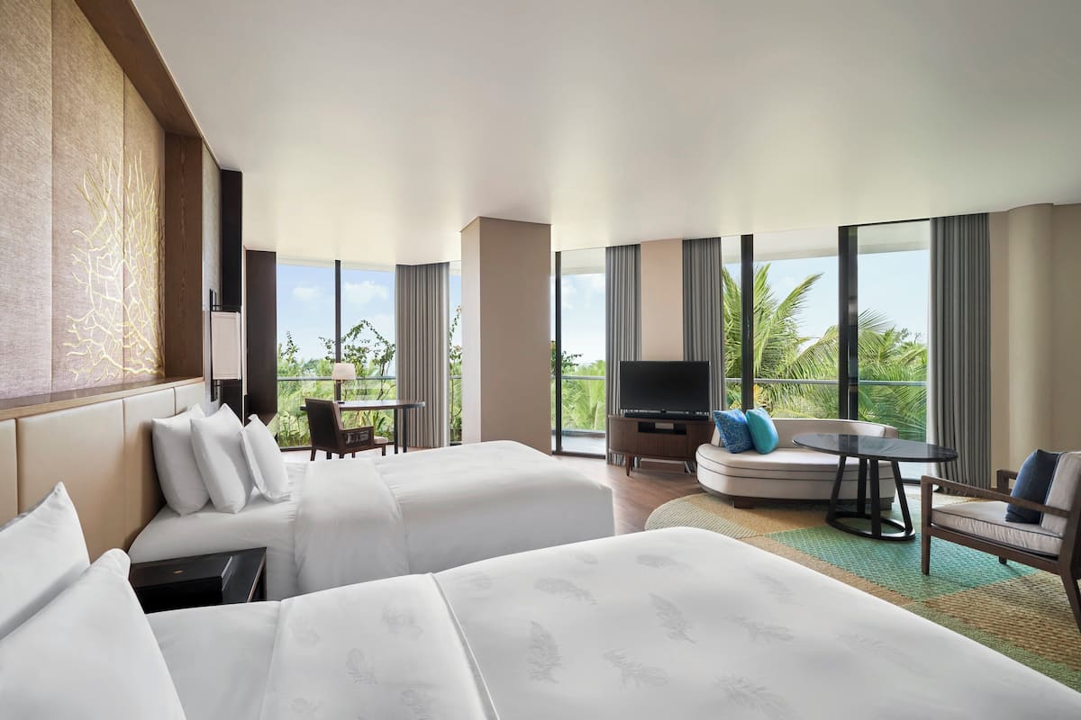 Habitación de hotel con dos camas, un sofá, un televisor y un balcón con vistas a palmeras.
