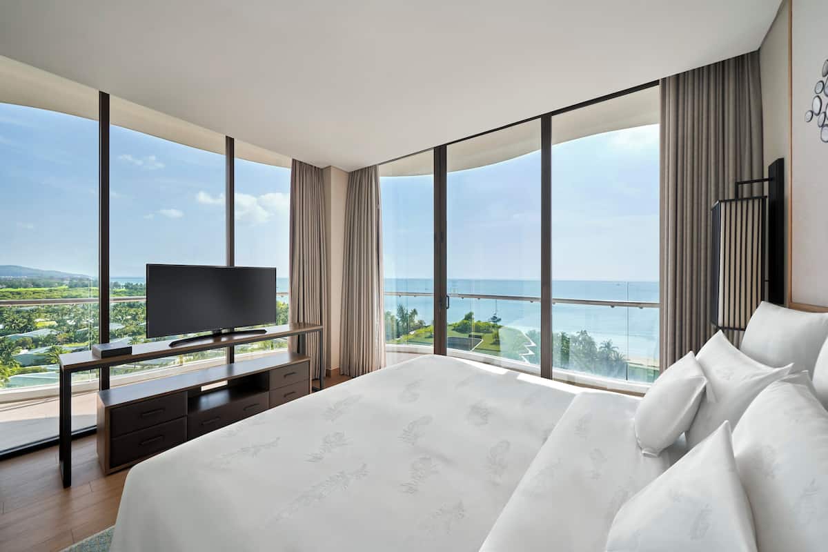 Une chambre d’hôtel moderne avec un grand lit, un téléviseur posé sur une console en bois et un balcon offrant une vue sur la ville.