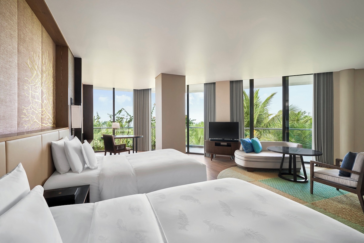 Habitación de hotel con dos camas, un sofá, un televisor y un balcón con vistas a palmeras.
