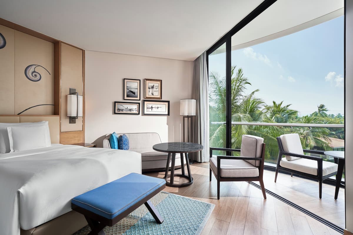 Une chambre d’hôtel avec un lit, un canapé, une petite table et des chaises. Des tableaux encadrés ornent le mur et, par la fenêtre, on peut admirer des palmiers.