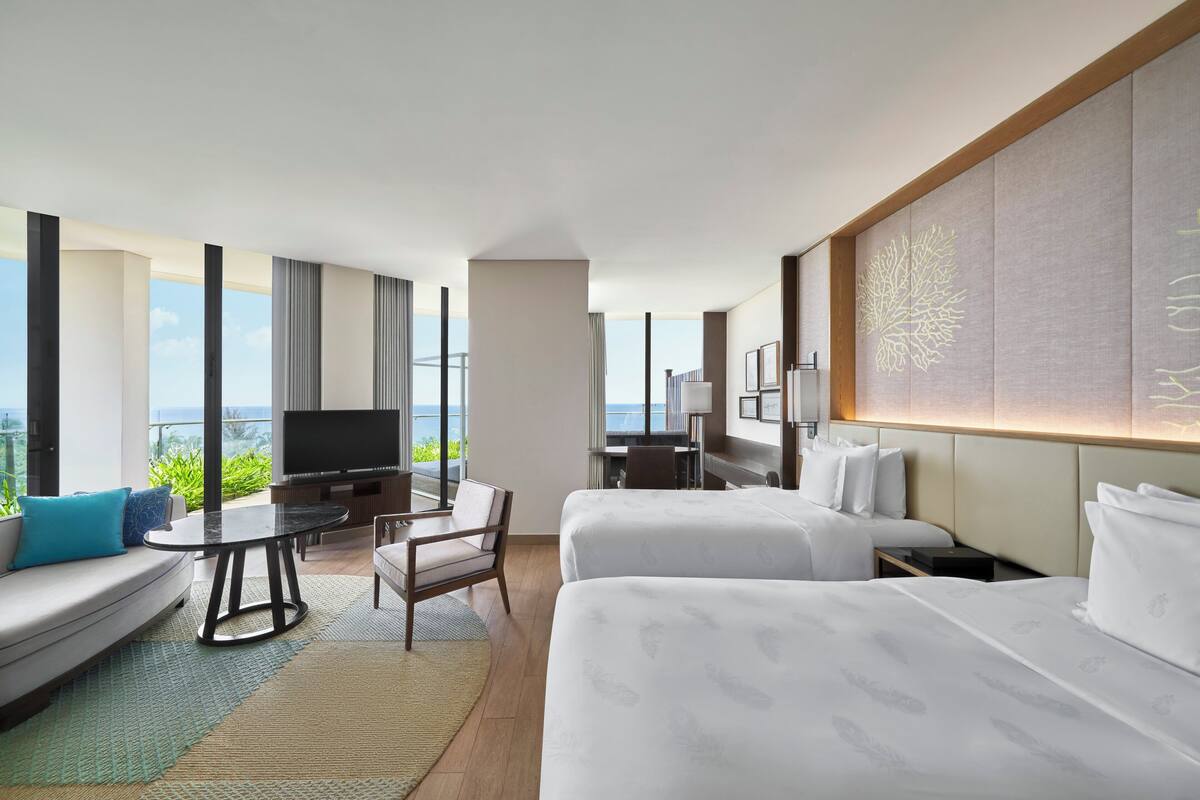 Une chambre d’hôtel moderne équipée de deux lits, d’un canapé, d’un bureau et d’une télévision. La chambre dispose de grandes fenêtres offrant une vue sur l’extérieur.