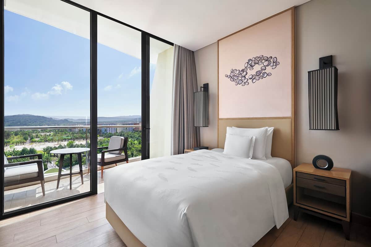 Une chambre d’hôtel avec un grand lit, une table de chevet, une chaise, une petite table et une vue sur le paysage urbain grâce à de grandes fenêtres.