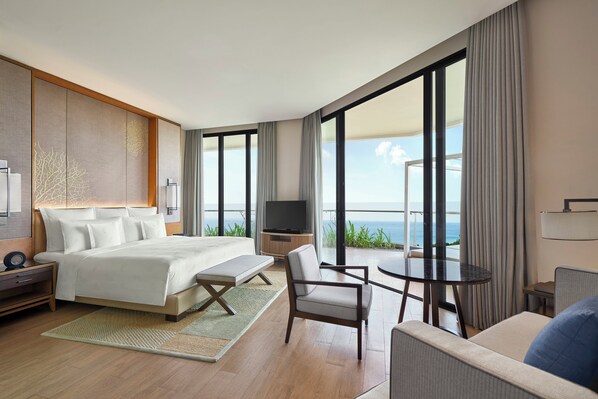 A modern hotel room with a large bed, a TV, a balcony with a view of the ocean, and a dining area.