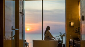 Una mujer sentada junto a un amplio ventanal con vista al océano al atardecer, en un interior moderno que incluye un sofá, una mesita y una planta en maceta.