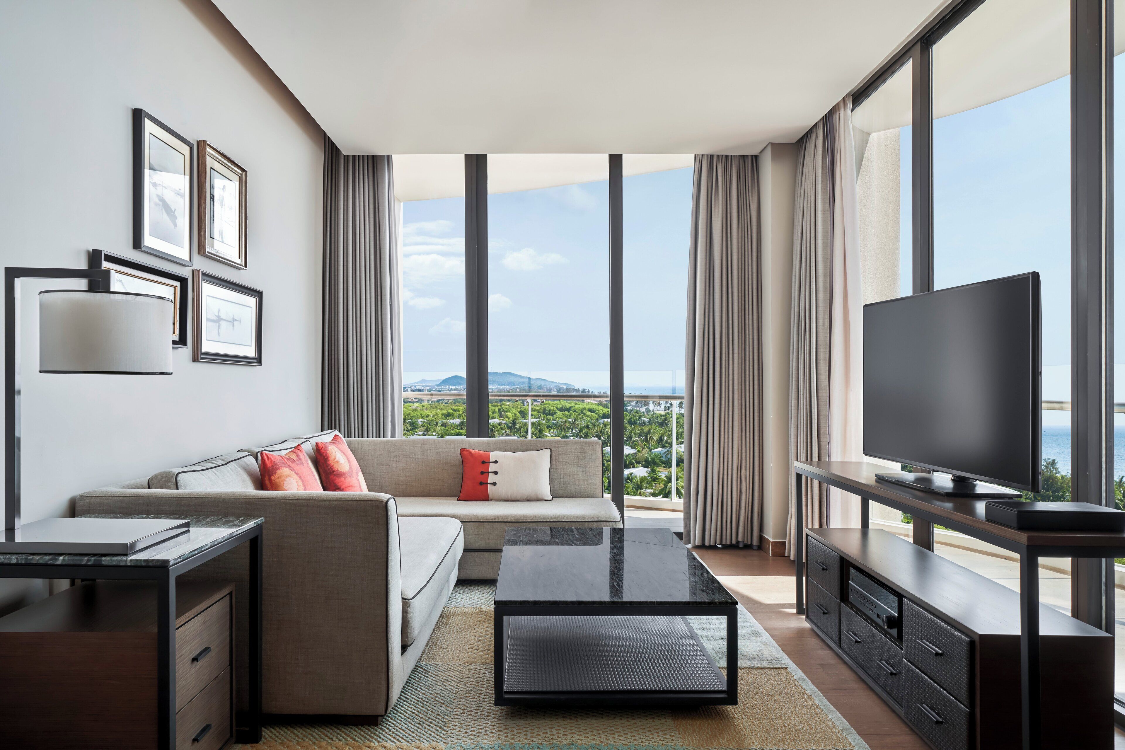 A modern hotel room with a sofa, a coffee table, a TV on a stand, and a desk with drawers. The room has large windows with a view of the city.