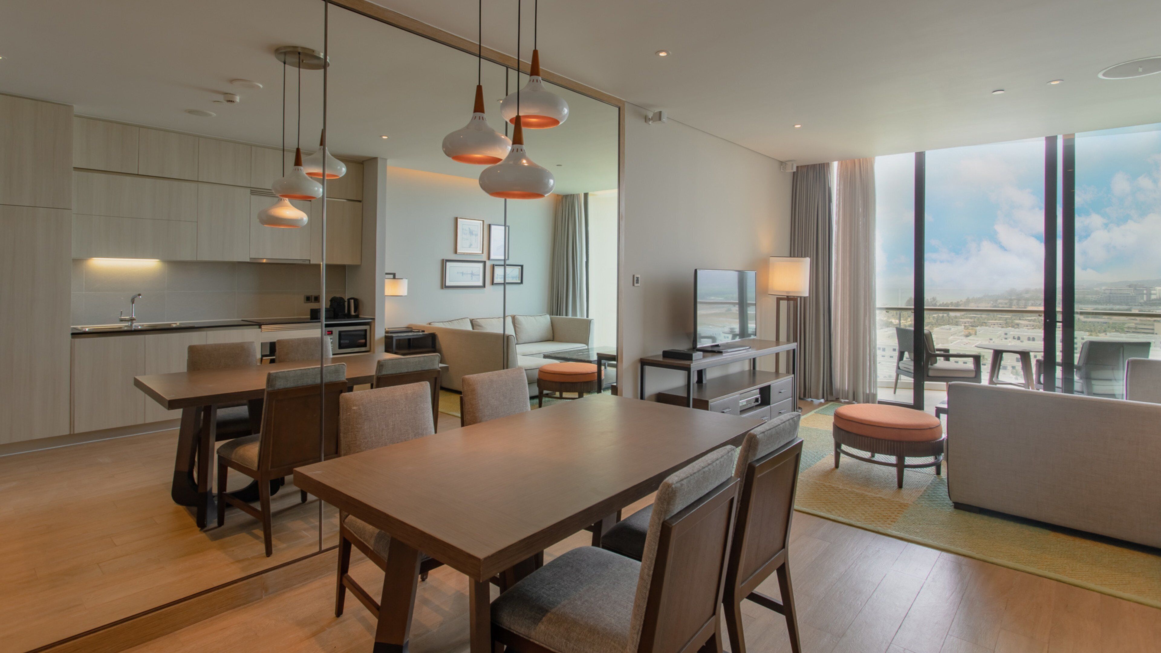 A modern hotel room with a dining area, kitchen, and a balcony with a city view.