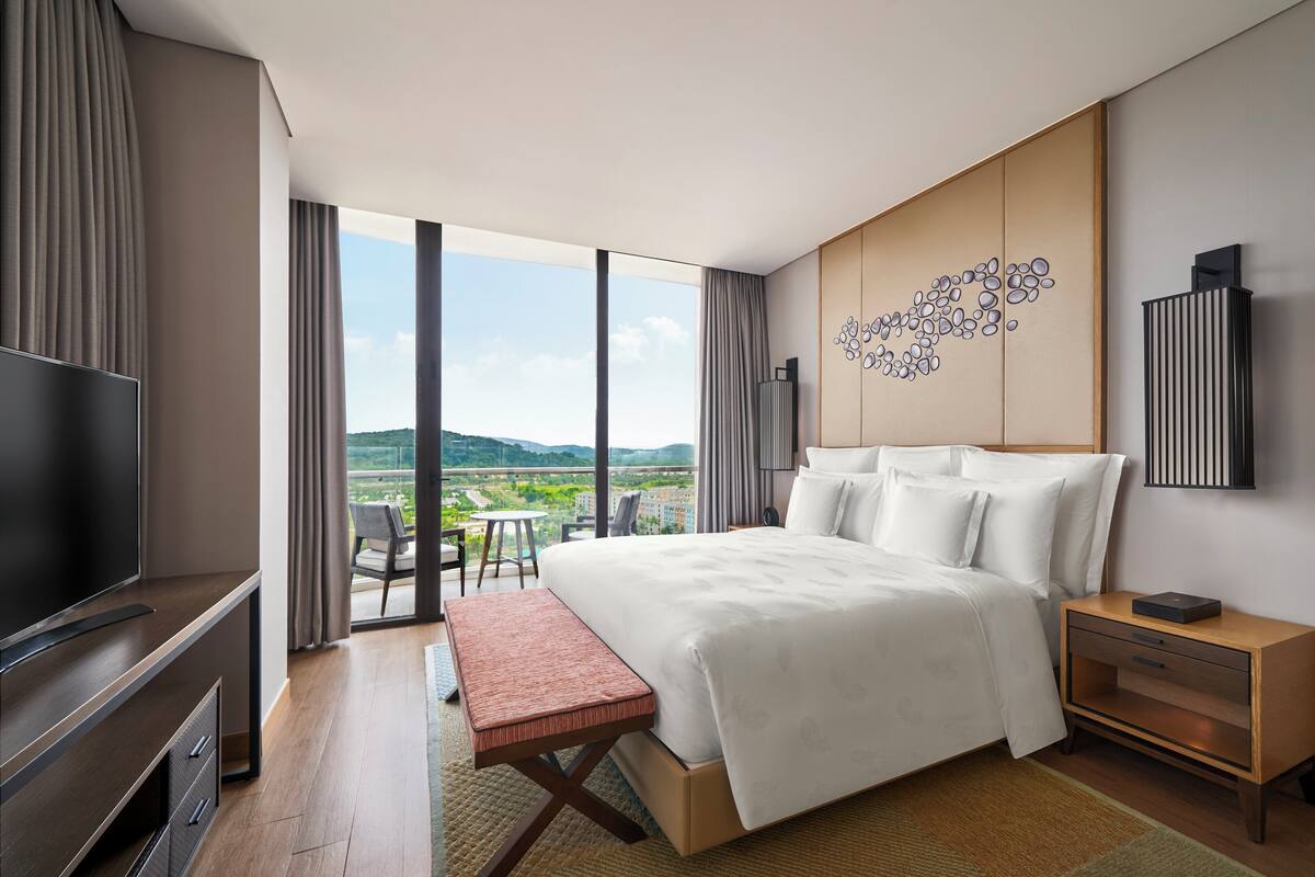 A modern hotel room with a large bed, a flat-screen TV, a wooden nightstand, and a view of the outdoors.