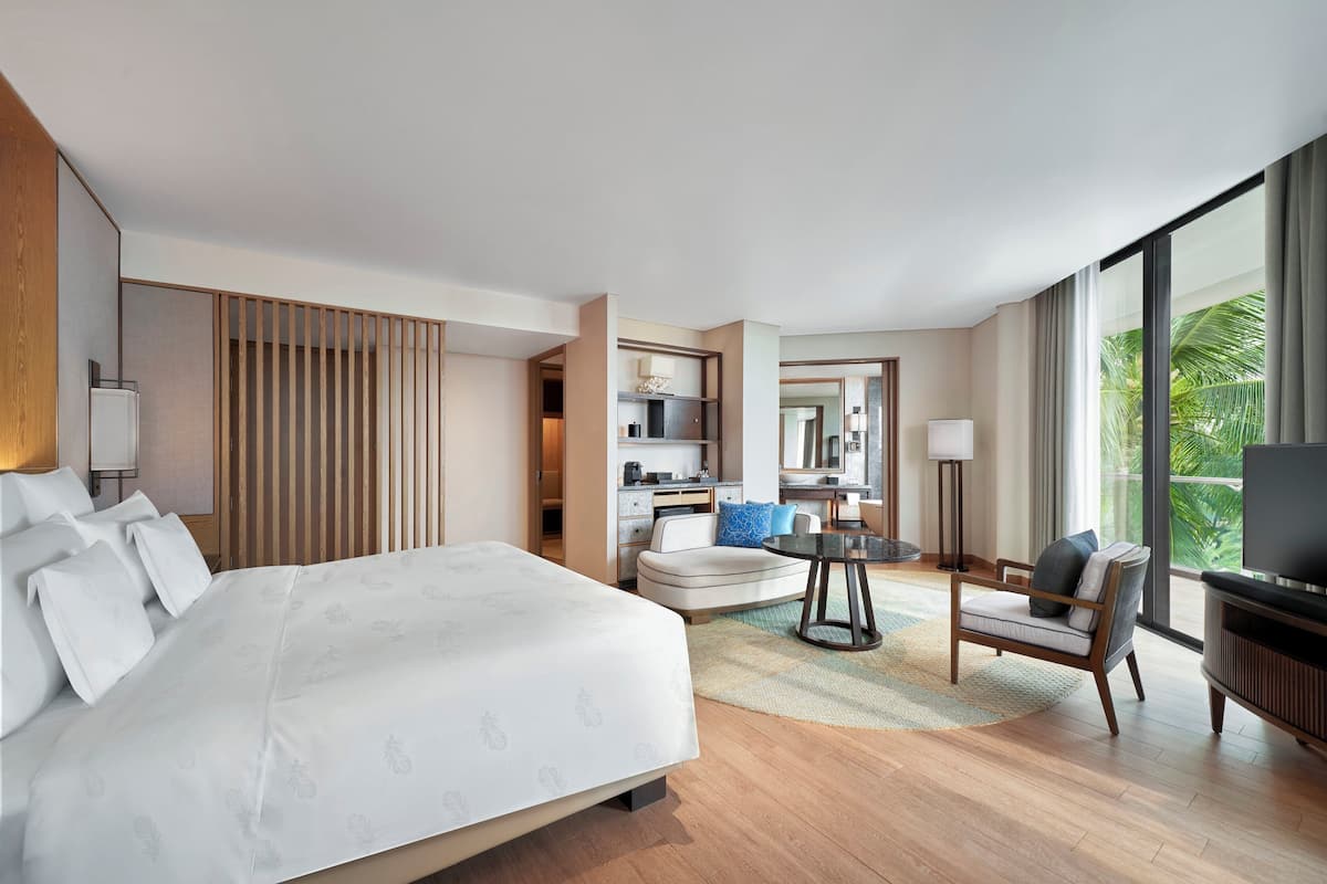 Une chambre d’hôtel avec un grand lit, un coin salon avec une table, une télévision et une vue sur la verdure environnante.