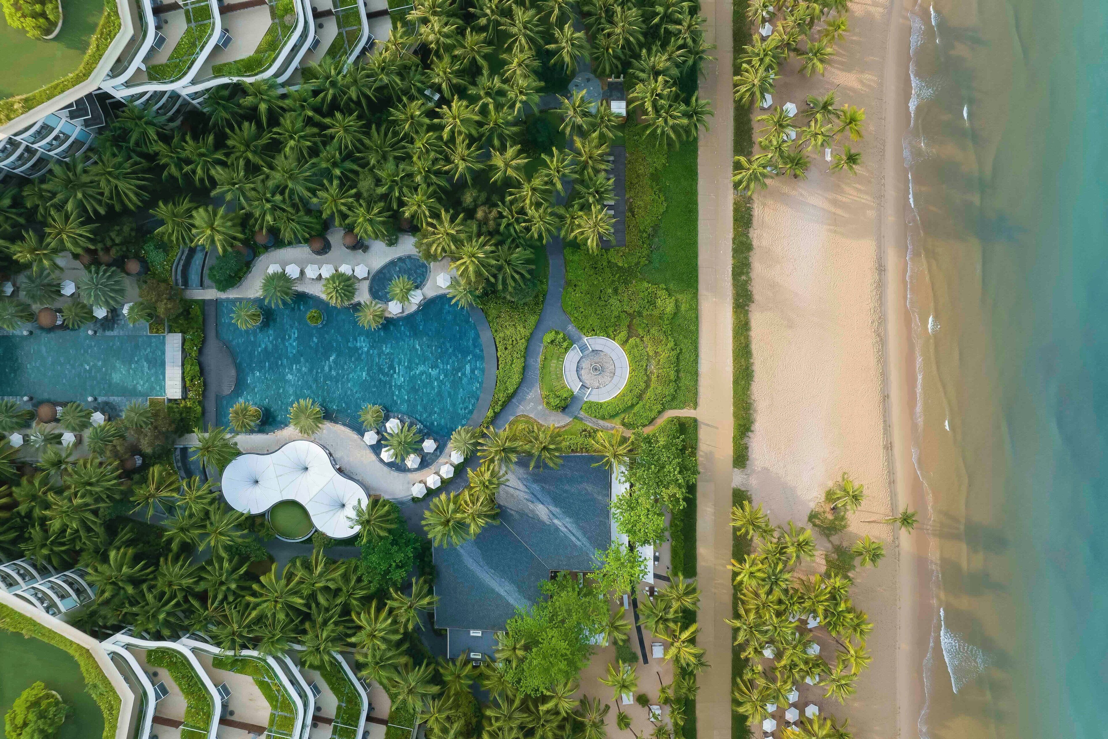 Vista aérea de un complejo turístico con piscina, palmeras y una playa.