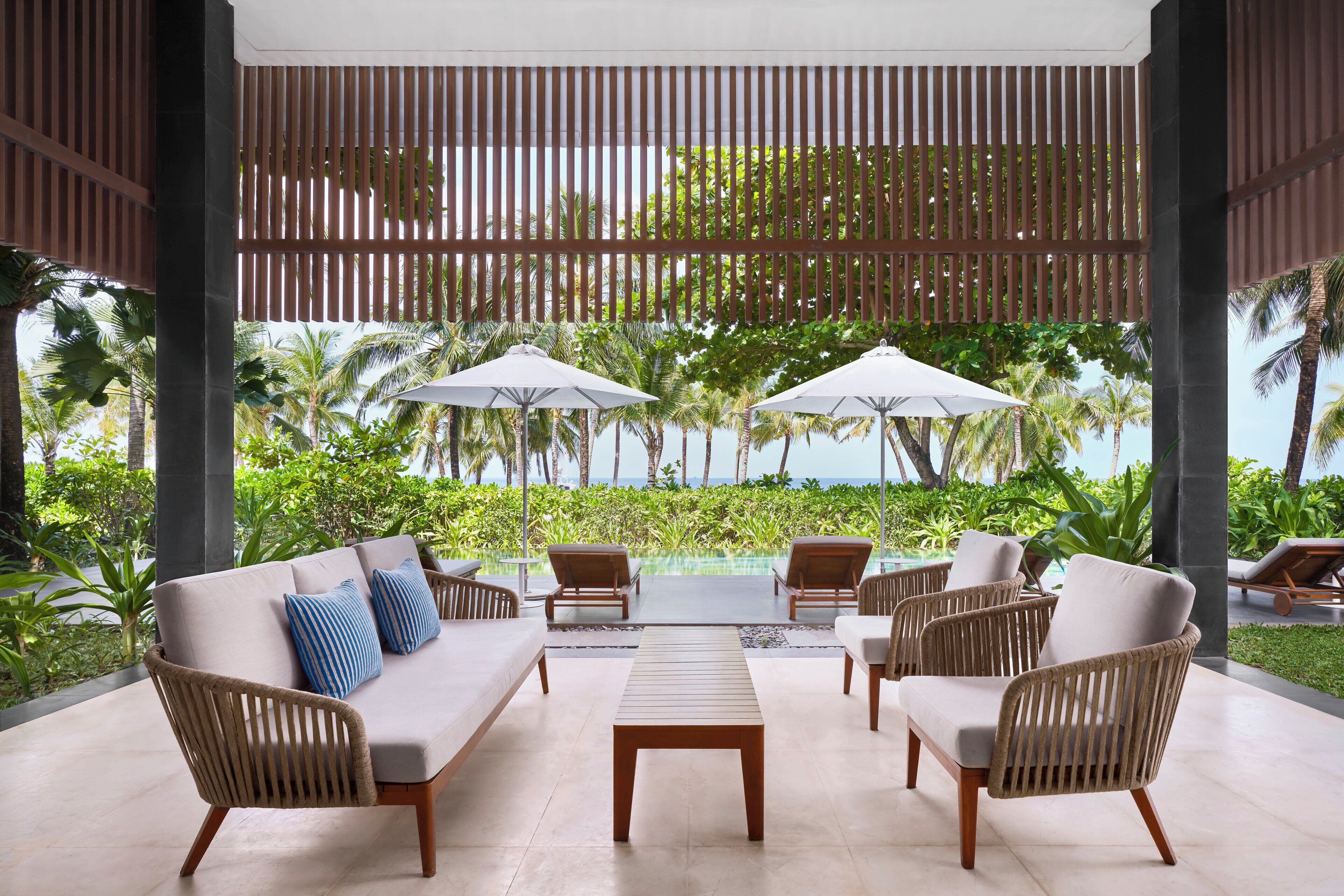 Un espace extérieur aménagé, abrité et doté de mobilier en osier, d’une table centrale et de parasols, offrant une vue sur une piscine et une végétation luxuriante.