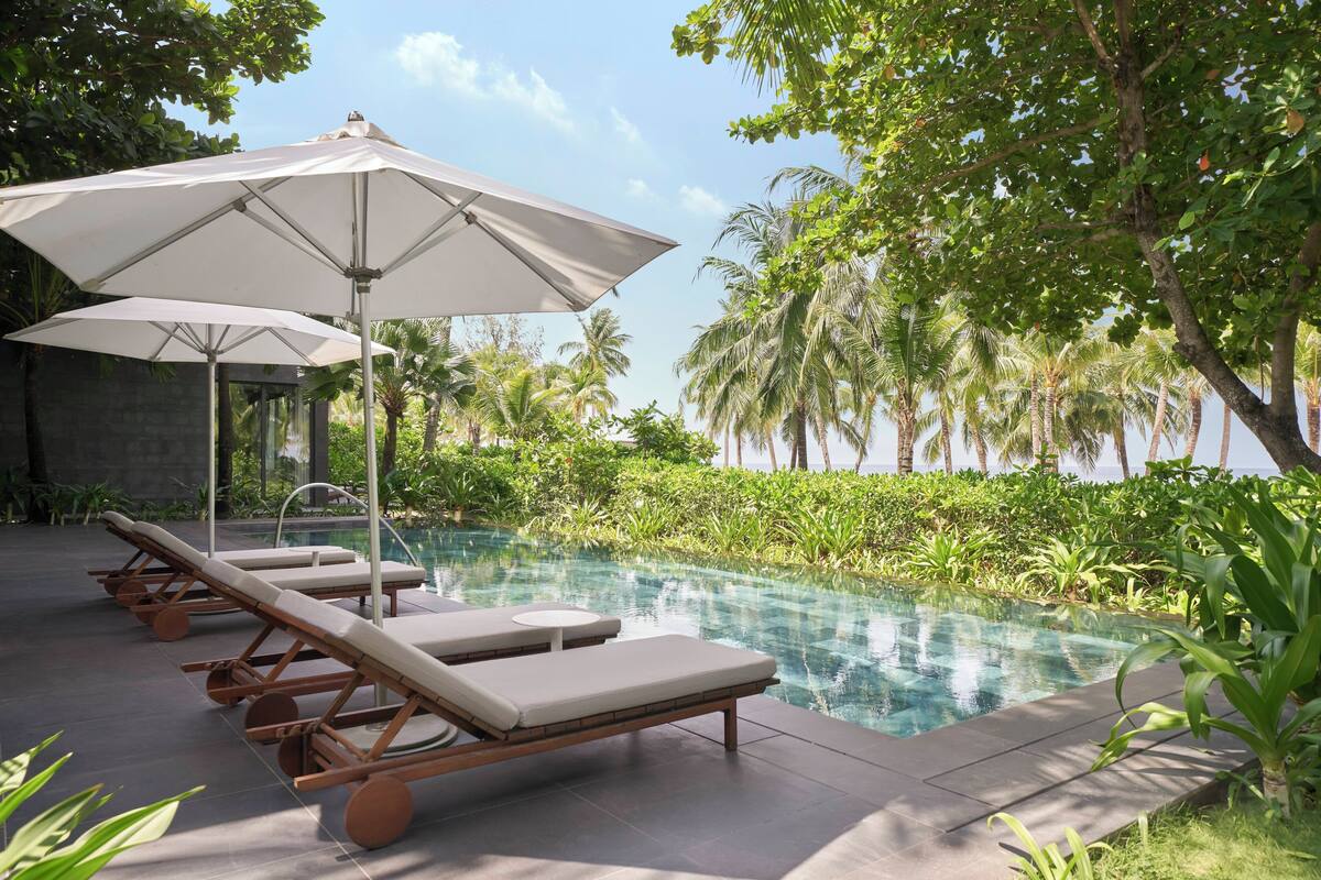 A poolside area with lounge chairs and umbrellas, surrounded by lush greenery and palm trees.