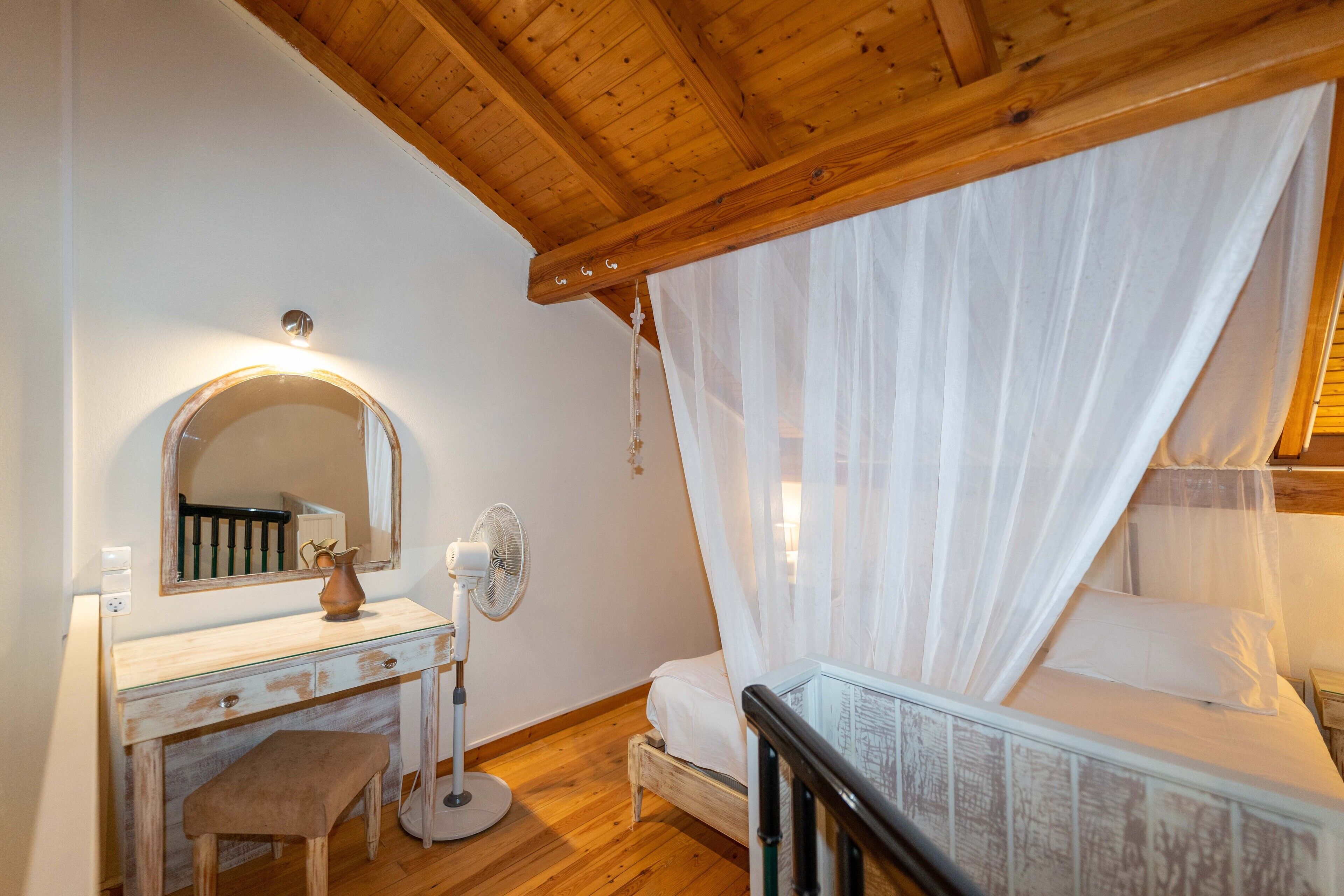 A bedroom with a wooden ceiling, a bed with white bedding, a bedside table with a fan, a mirror, and a bench.