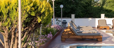 A poolside area with lounge chairs, a pool, and a pathway lined with trees and flowers.