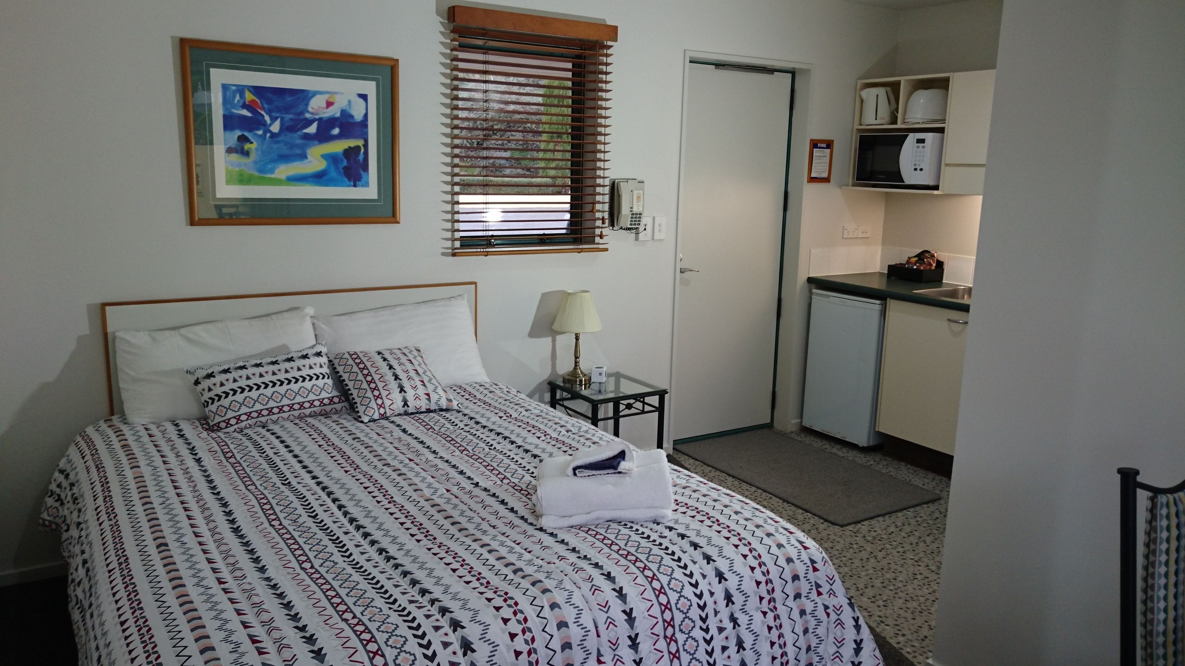 A bedroom with a bed, a bedside table with a lamp, a painting on the wall, and a kitchenette with a microwave and coffee maker.
