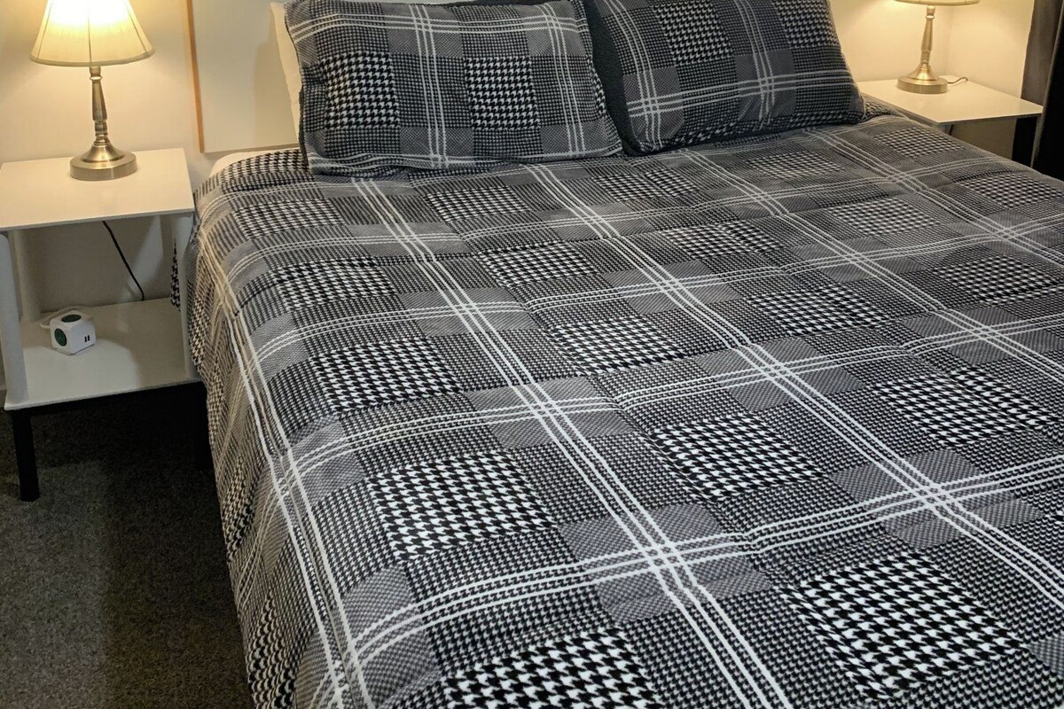 A bed with a checkered bedspread, two bedside lamps, a framed painting above the bed, and a small bedside table with a lamp.