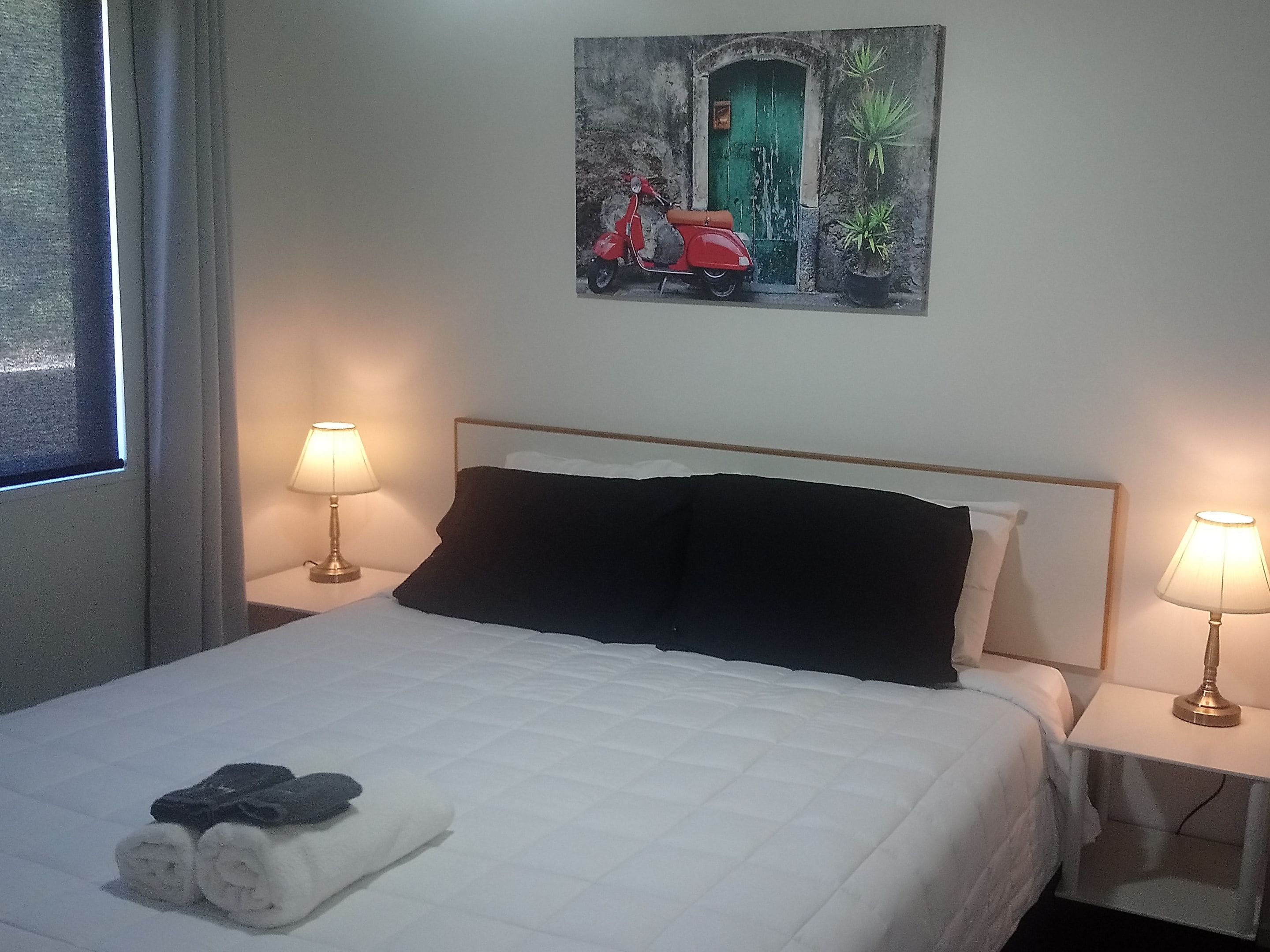 A hotel room with a bed, two bedside lamps, a painting on the wall, and a window with curtains.