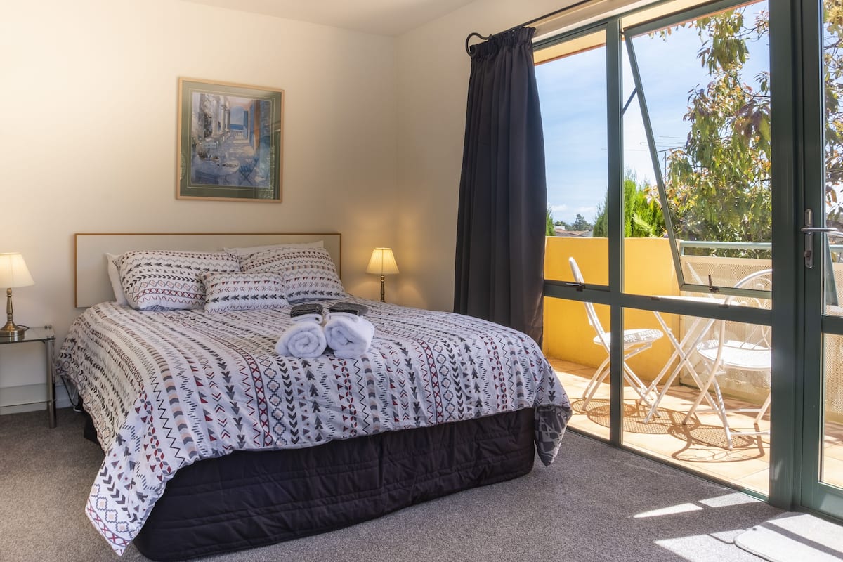 A bedroom with a bed, a painting, a window with curtains, and a balcony with chairs.