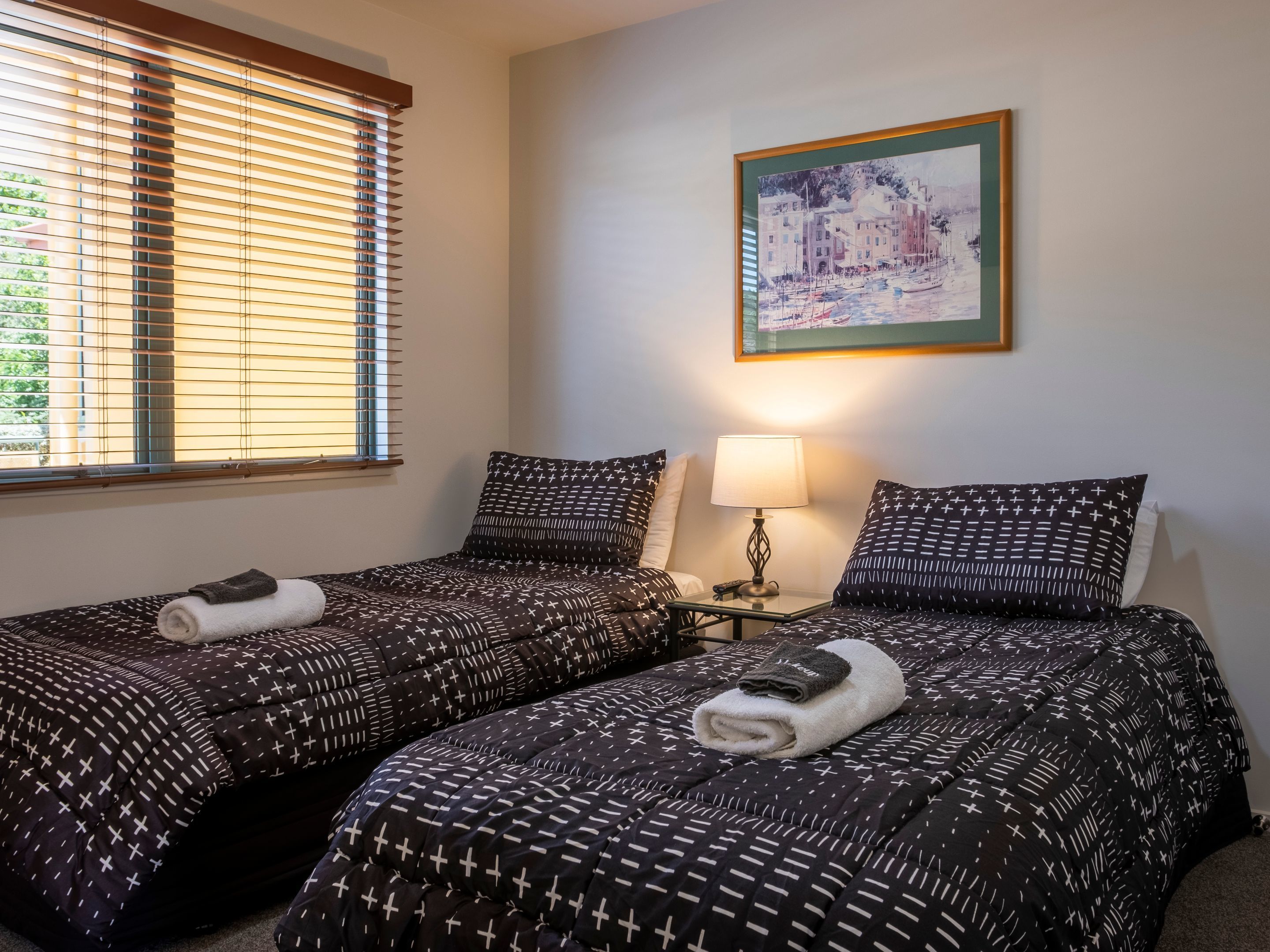 Habitación de hotel con dos camas, cada una con ropa de cama estampada y una toalla enrollada encima. Una mesita de noche con una lámpara y un cuadro en la pared.