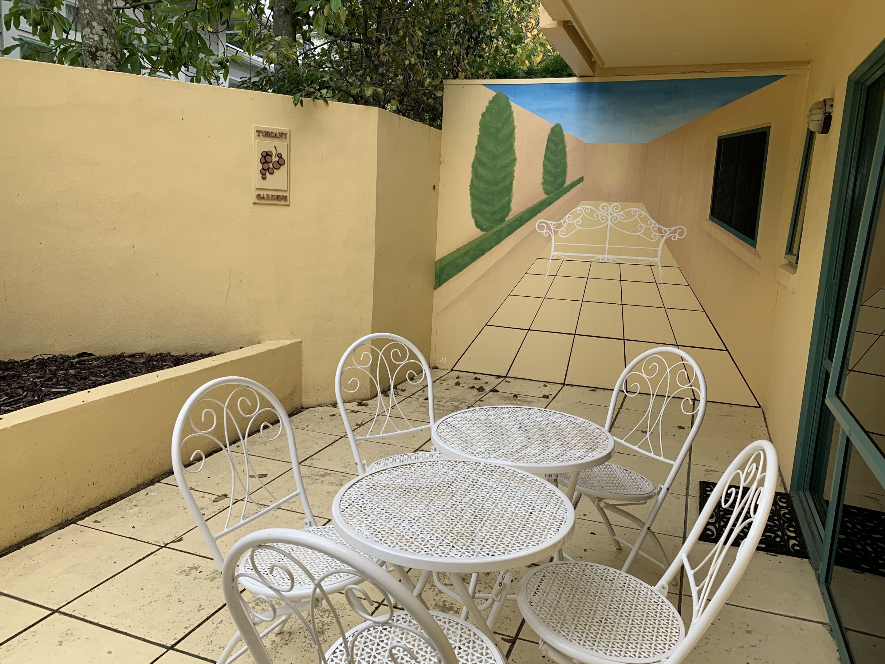 Un patio con sillas y mesas de metal blanco, suelo de baldosas y un mural en la pared.