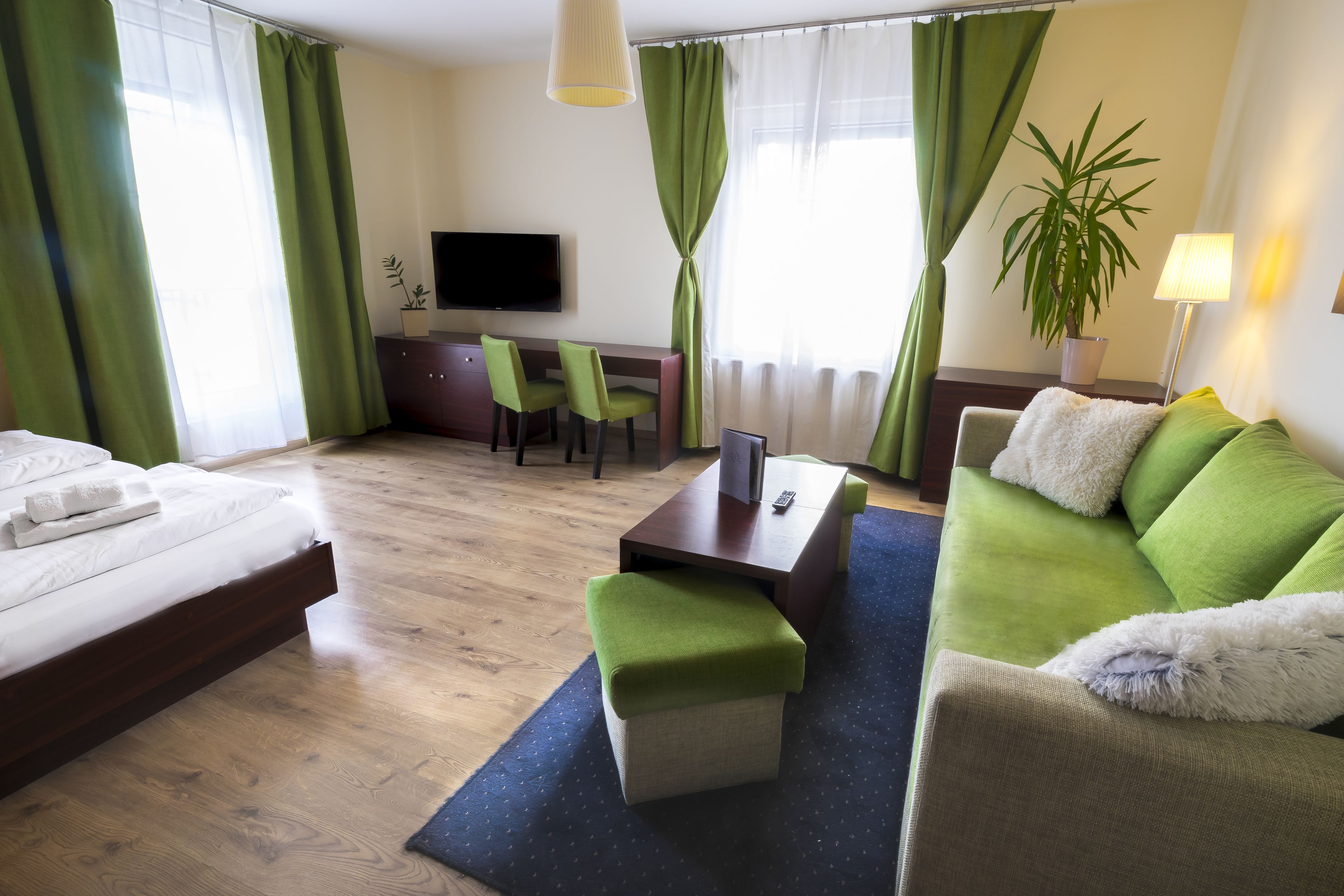 Habitación de hotel con cama, sofá verde, una mesita, televisor y planta en maceta.
