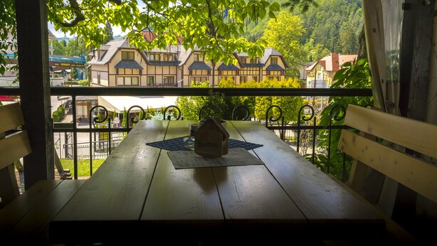 Una mesa de madera con un mantel individual y un pequeño objeto con forma de casa, situada en un balcón con barandillas de metal y vistas a edificios tradicionales y follaje.
