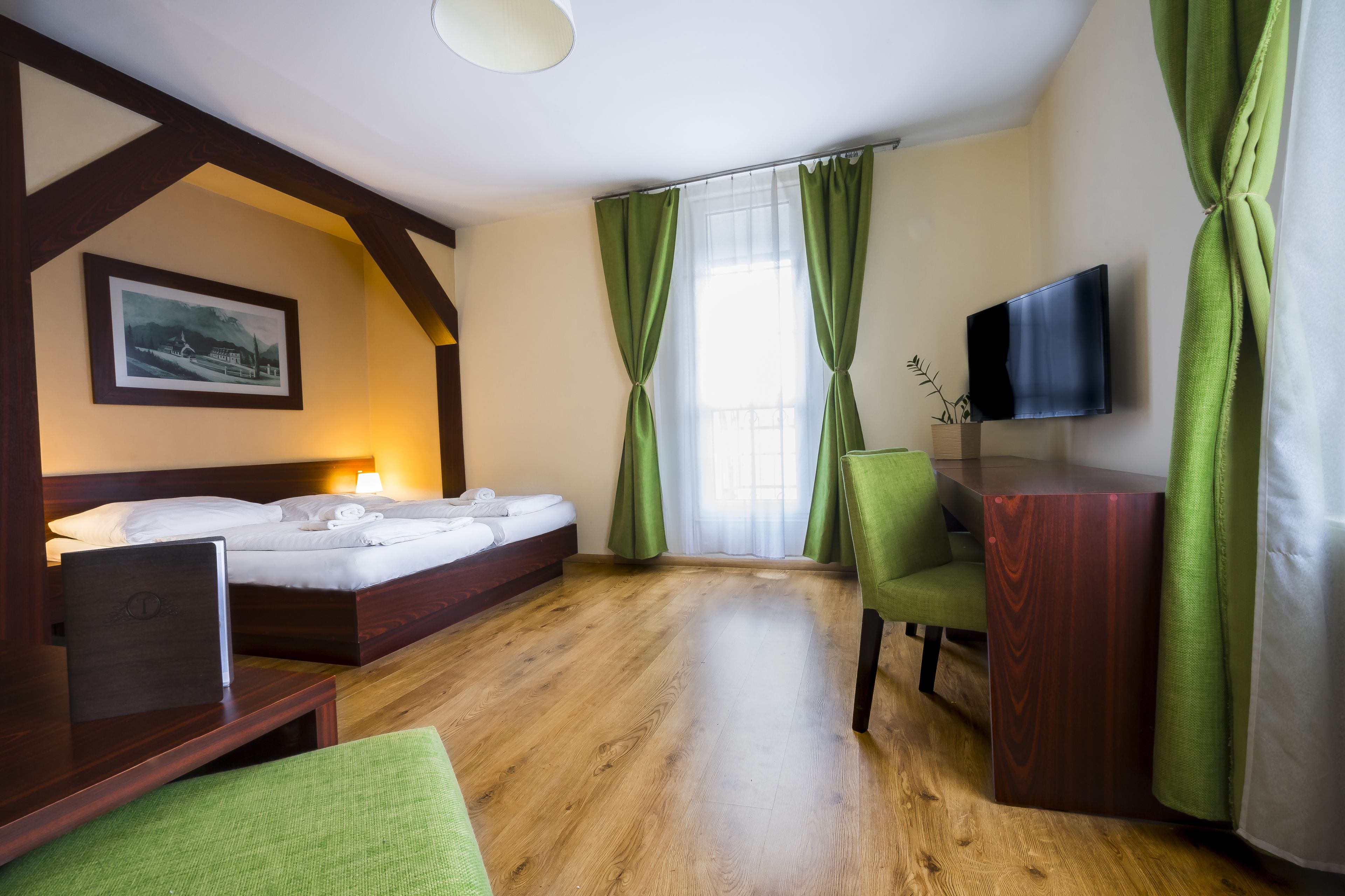 A hotel room with a wooden floor, a bed with white linens, a desk with a laptop, a TV on the wall, and green curtains.