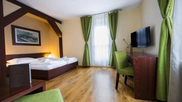 A hotel room with a wooden floor, a bed with white linens, a desk with a laptop, a TV on the wall, and green curtains.