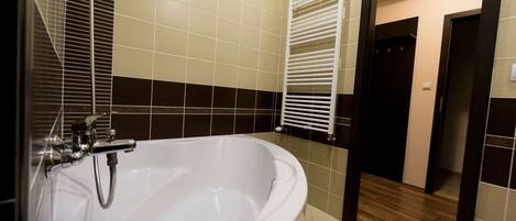 Un baño con una bañera blanca grande, azulejos color beige y marrón oscuro, un perchero para toallas y suelo de madera.