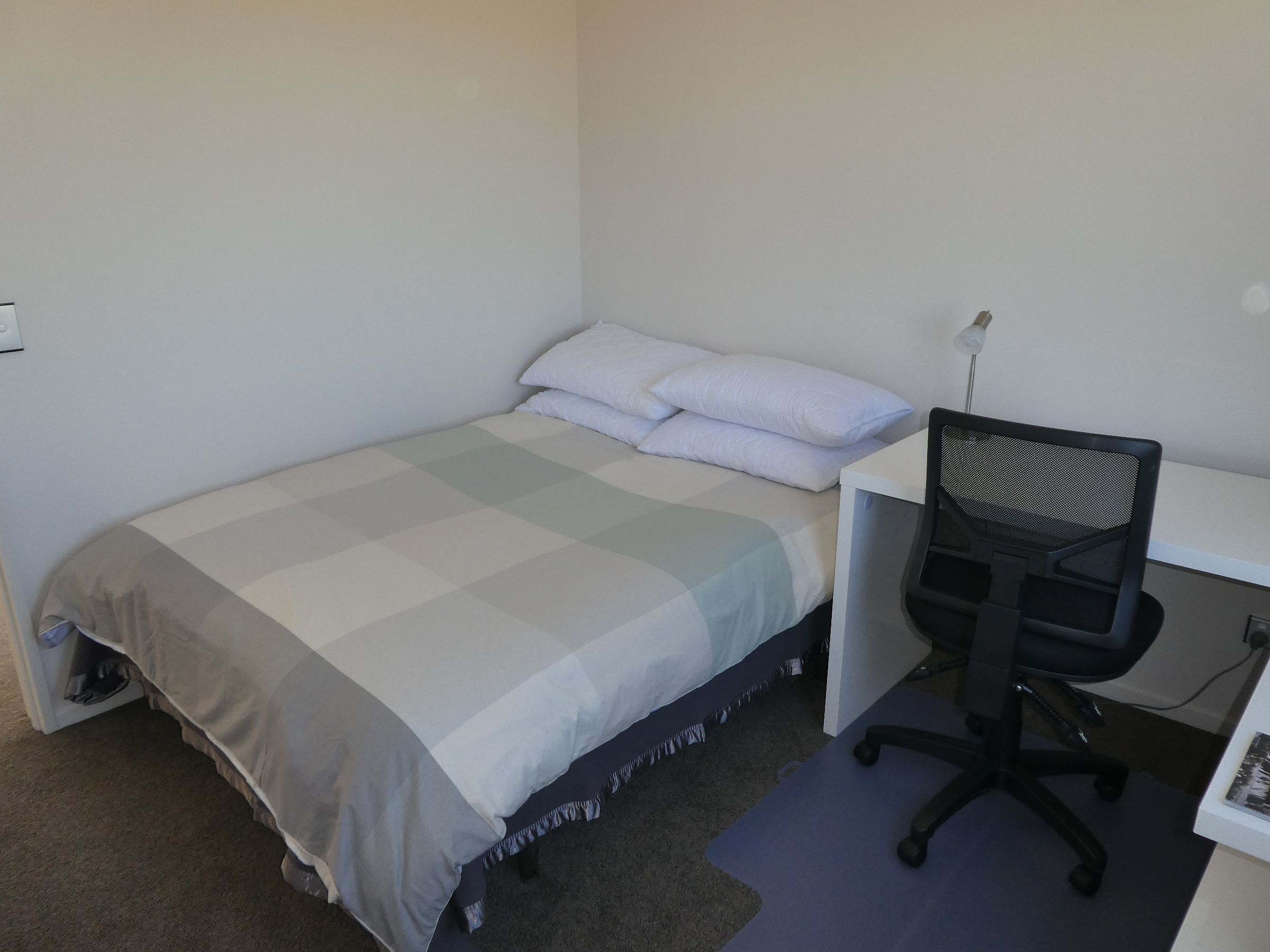 A small, single-bed room with a desk and chair, a power outlet, and a carpeted floor.