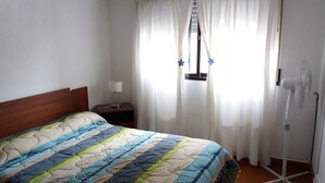 A neatly made bed with a colorful quilt, a bedside table with a lamp, a window with curtains, and a standing fan. - Como en casa (Rosario)