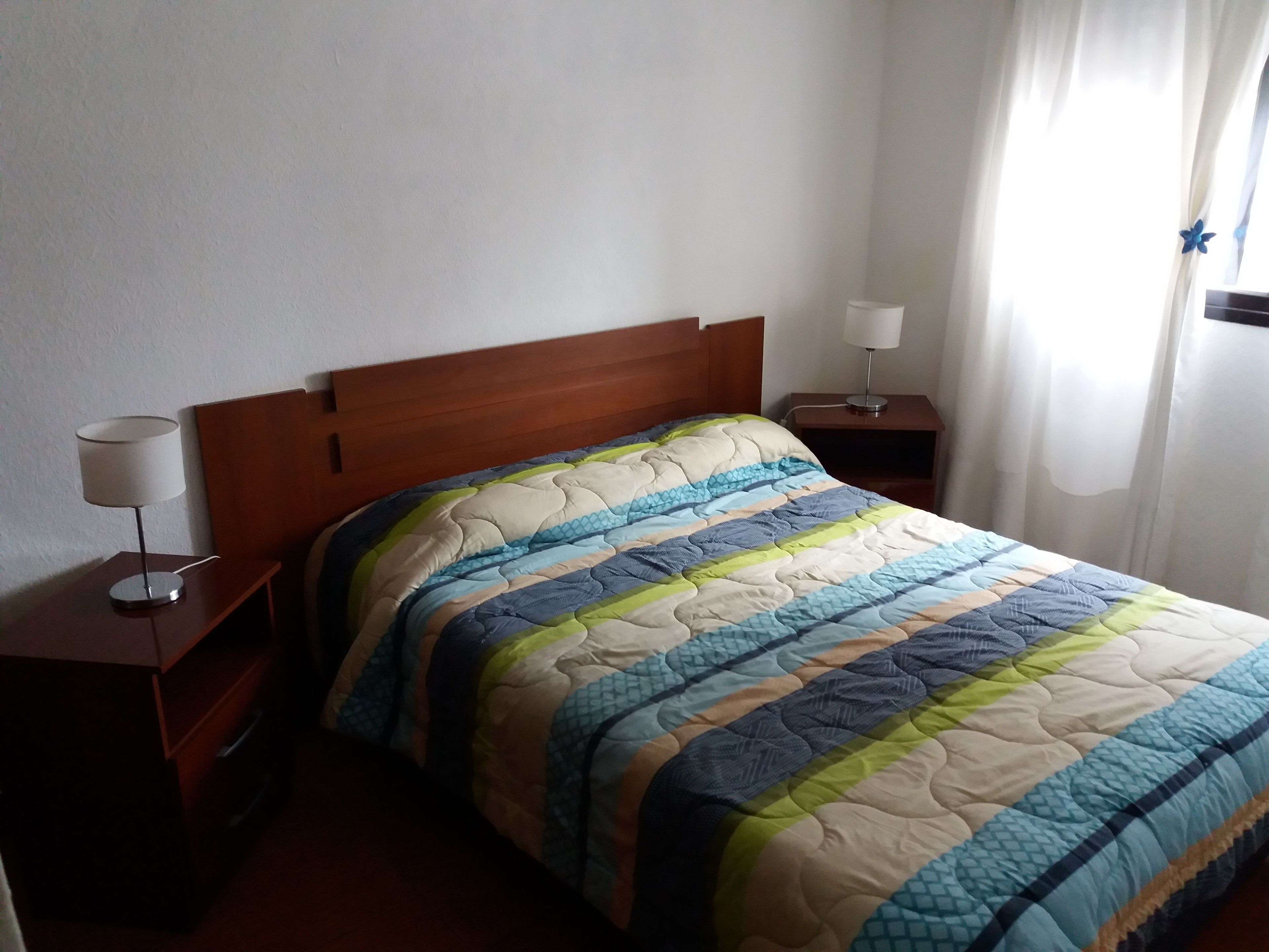 Una cama bien tendida con un edredón a rayas, dos mesitas de noche con lámparas y una ventana con cortinas.