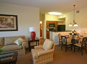 Una sala de estar con sofá, sillón y una mesa de comedor con sillas. Cuenta con una zona de cocina con armarios y microondas.